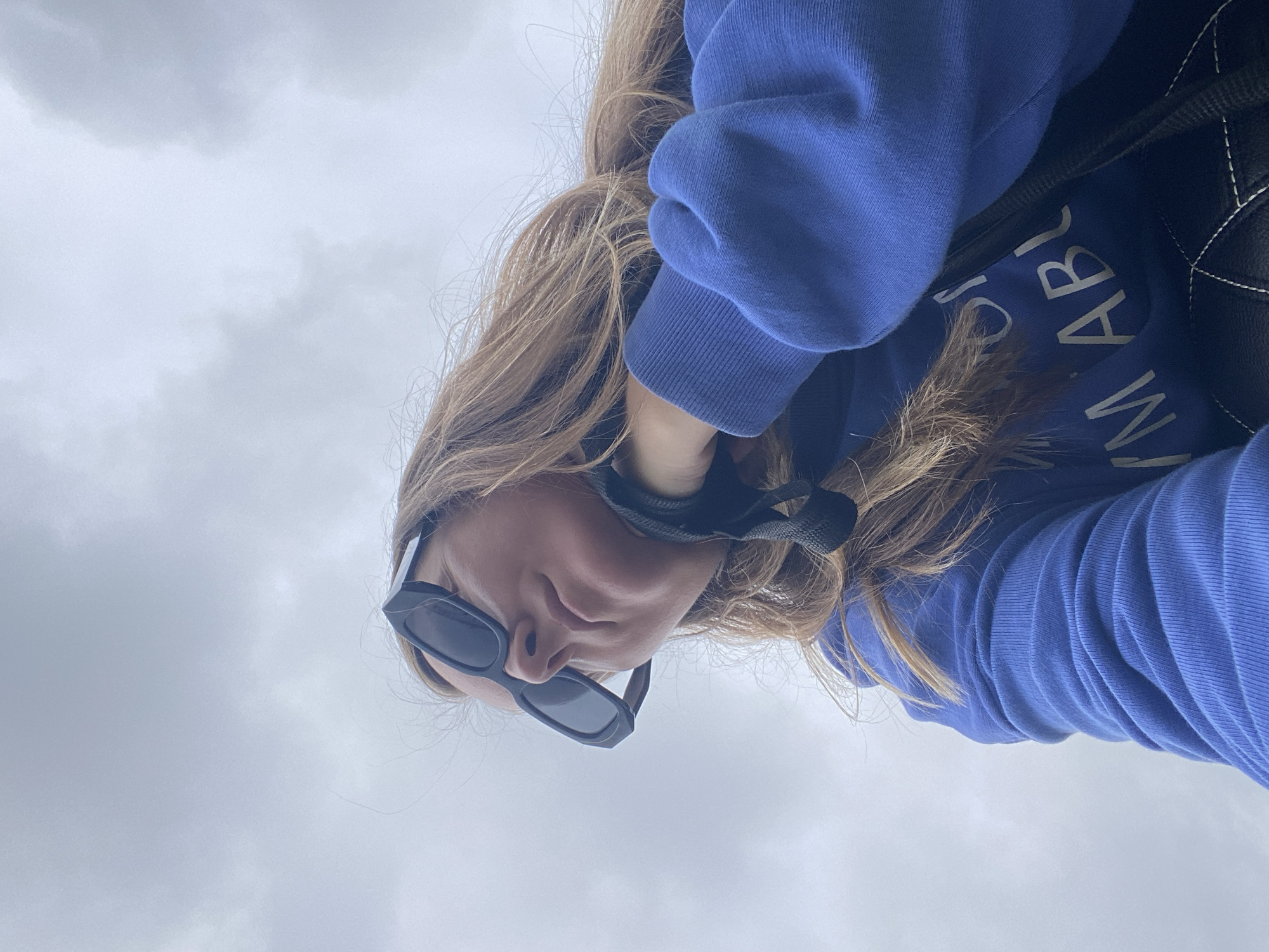 The image shows a person from a low angle, looking up at the sky, which is cloudy. The person is wearing a blue sweatshirt and black sunglasses. Their hair is long and blonde, and they are holding one hand up to their neck, with a black strap visible. Part of the sweatshirt has white text on it, with the visible words being "M ABLE." The background is entirely composed of the sky. There is no discernible urban environment or any specific location within Noviy, Russia visible in the image.