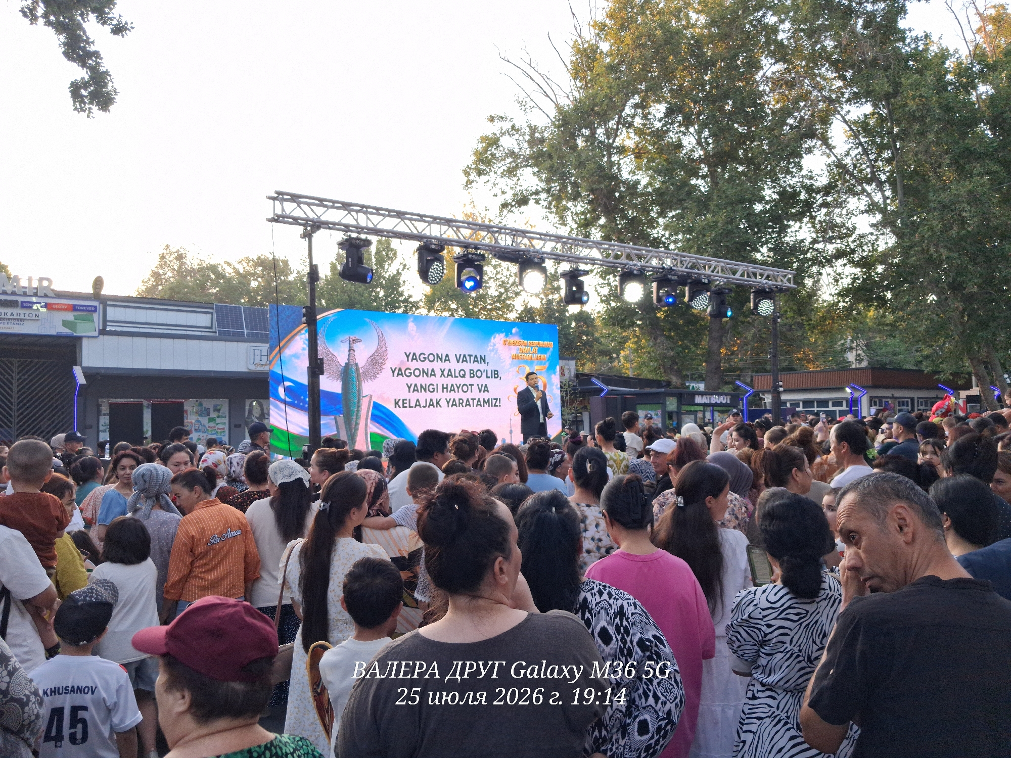 The image depicts an outdoor gathering of people, predominantly women and children, in front of a large LED screen displaying a congratulatory message in Uzbek, along with an image of the Uzbek national emblem. The text on the screen reads, "YAGONА VATAN, YAGONА XALQ BO'LIB, YANGI HAYOT VA KELAJAK YARATAMIZ," which translates to "One Homeland, One People, we will create a new life and future." A man in a suit is standing on the stage in front of the screen, appearing to be addressing the crowd. The scene is set in an urban environment, with trees lining the background. There are stage lights suspended above the screen, indicating a performance or event is taking place. The crowd is dense, with individuals of various ages dressed in casual attire. Some people are holding phones, possibly to record the event. The overall atmosphere appears festive and communal. The timestamp at the bottom of the image indicates the photo was taken on July 25, 2026, at 19:14. This event is identified as taking place in Chirchik, Uzbekistan.