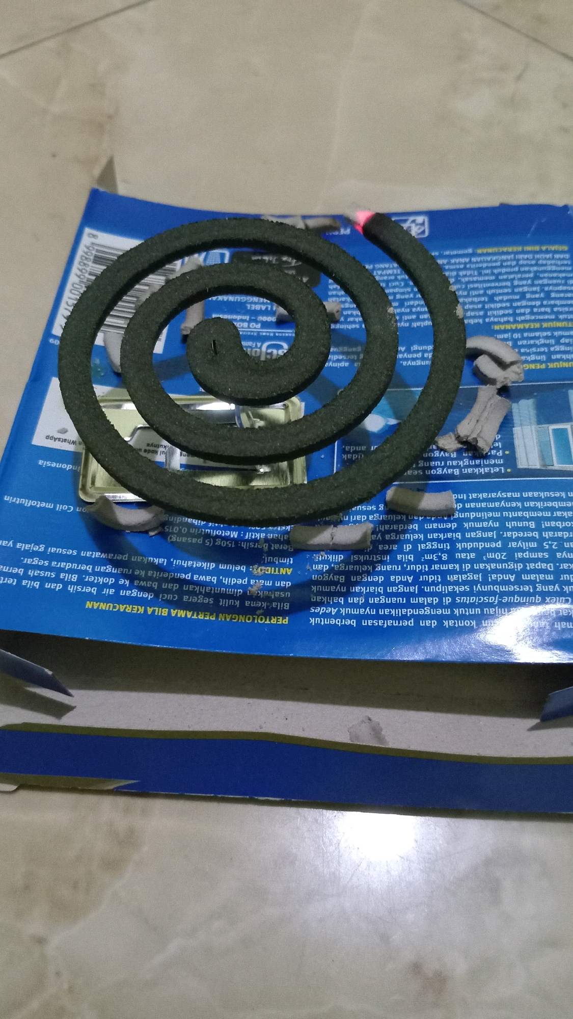 This is a close-up, overhead shot of a mosquito coil, partially burned, resting on a blue and white cardboard box with Indonesian text. The mosquito coil is dark green and coiled in a spiral shape, with the end glowing red, indicating it is lit. The cardboard box appears to be packaging for the mosquito coil. The text on the box is in Indonesian and includes labels such as "LABEL," "PD BOX," "A Family of Baygon," and warnings and instructions. The surface beneath the box appears to be a marble or tiled countertop. The overall scene suggests an indoor environment where mosquito repellent is being used. There are no people visible in the image. The location is identified as Randuagung, Indonesia, based on the text on the box.