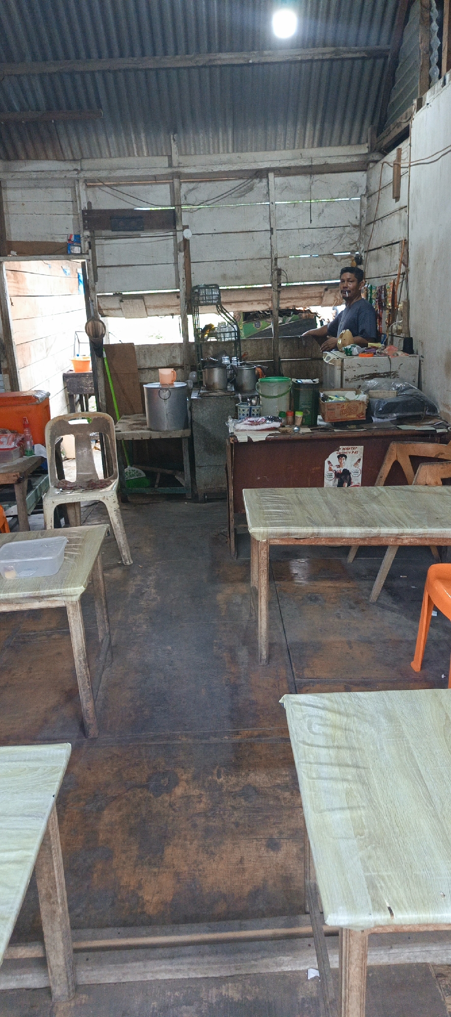 The image depicts the interior of a humble eatery in Garot, Indonesia. A man, appearing to be the proprietor, stands behind a counter that holds various kitchen supplies and containers. He is wearing a dark t-shirt and has a cigarette in his mouth. The space is characterized by its rustic construction, with a corrugated metal roof and wooden walls and floors. Several tables and chairs are arranged in the dining area, some covered with light-colored plastic. Various containers, including buckets and large pots, are visible in the background, suggesting food preparation is ongoing. The overall impression is that of a simple, functional establishment catering to locals.