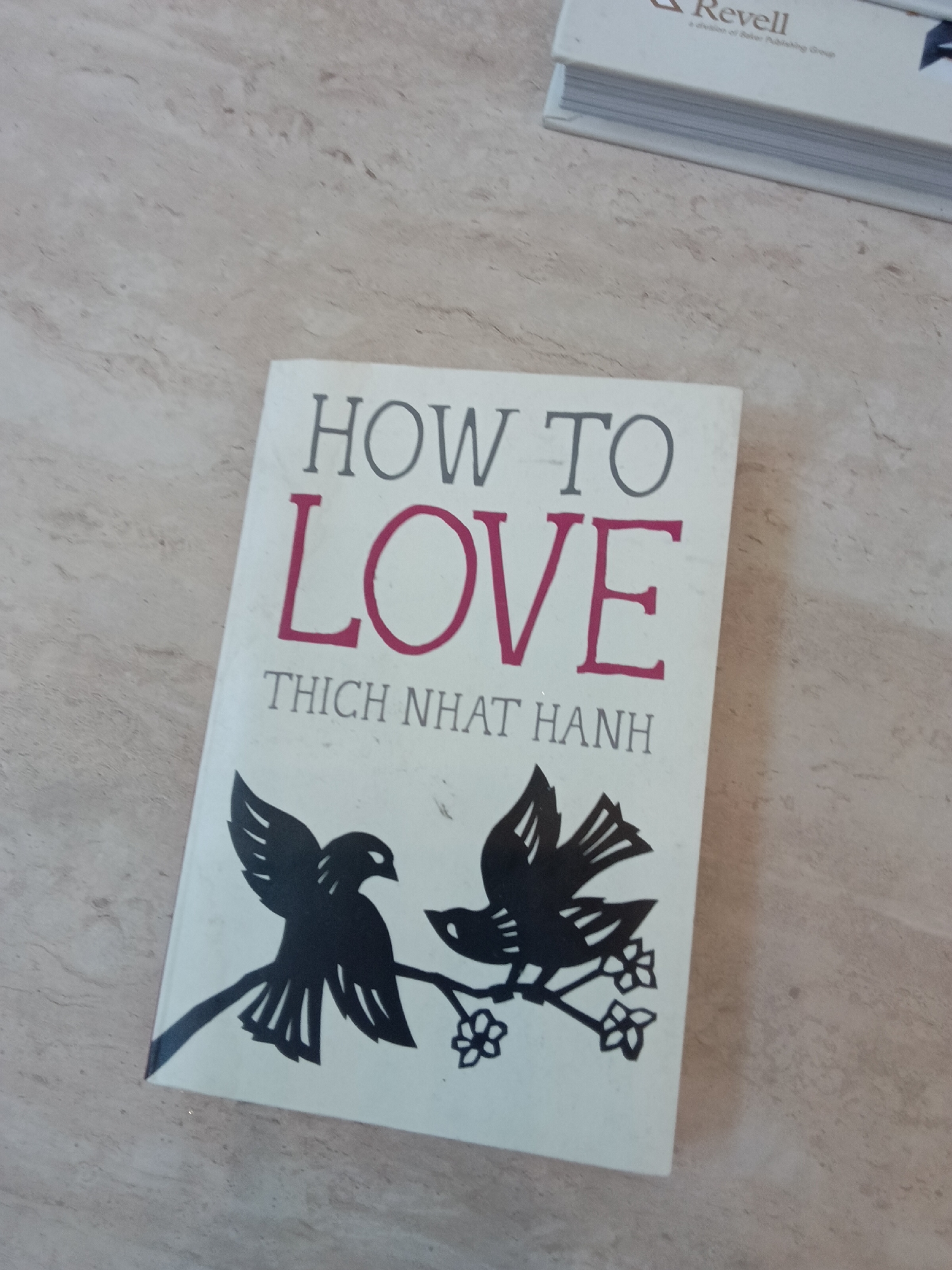 The image shows a book titled "How to Love" by Thich Nhat Hanh. The title is printed in gray and red. Below the title is the author's name, "Thich Nhat Hanh." The cover also features a black and white illustration of two birds on a branch. The book is lying on a surface that appears to be a countertop or table. In the upper right corner, there is a glimpse of another book. The scene appears indoors.