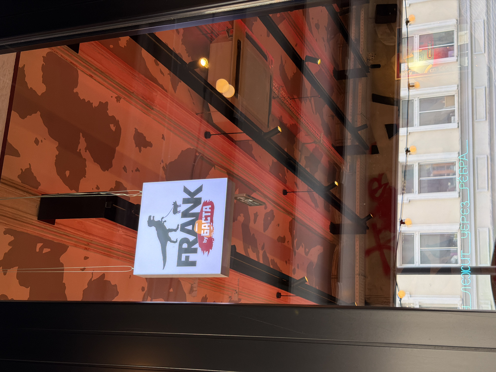 The image shows a sign for "FRANK by BASTA" reflected in a window. The sign features a silhouette of a dinosaur on a leash with a small animal. The ceiling above is painted with an orange and brown camouflage pattern. The reflection also shows a street scene with buildings and windows, as well as string lights and other lighting fixtures. There is a neon sign visible in the reflection with blue Cyrillic text that appears to say "ЛЕЖИТ ЧЕРЕЗ РЕБРА". This phrase, translated to English, means "lies through the ribs," which is likely a reference to a specific dish or menu item. The overall impression is that of a restaurant or food establishment. The context suggests the image was taken in Moscow, Russia, due to the Cyrillic text and common architectural styles.