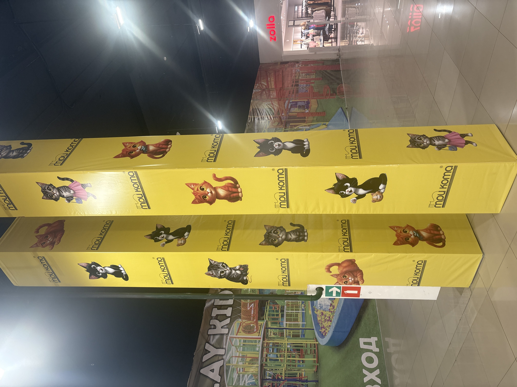 Here's a matter-of-fact description of the scene:
The photograph shows an indoor scene, likely within a shopping mall in an unknown city. In the foreground are three yellow rectangular pillars stacked on top of each other, each adorned with cartoon illustrations of cats and the brand name "mpu koma". These pillars appear to be an advertisement or a display. Below the pillars is a sign with text that partially reads "AY KID" and "ВХОД". There is also a graphic of a play area.
In the background, partially visible through a glass partition, is a store named "zolla". The floor appears to be a light colored tile. The lighting is bright.