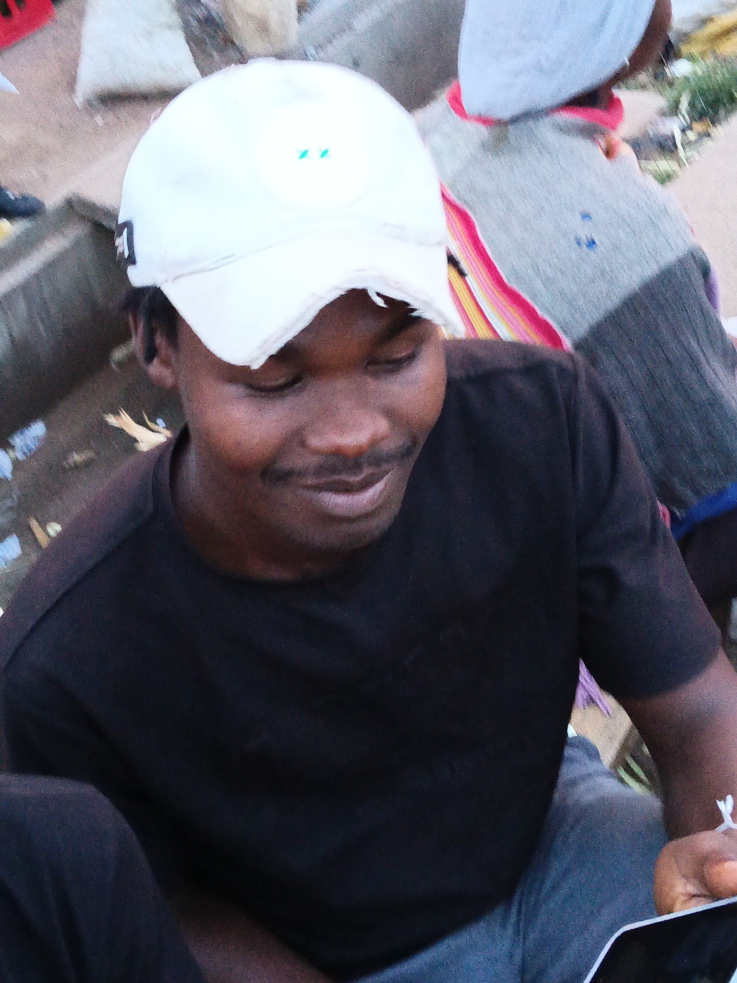 In Bukuru, Nigeria, a young Black male wearing a black t-shirt and a white baseball cap is the central figure. He is looking down, with his eyes closed, and a slight smile on his face. His mouth is slightly open, revealing his teeth, and he has a mustache. He is wearing dreadlocks that are visible under his cap.
Behind him, another person, likely a child, is wearing a grey sweater with colorful stripes. Their face is not clearly visible, but they are wearing a light blue beanie. The background suggests an outdoor setting with some debris on the ground, including plastic bags and what appears to be a broken piece of wood. The overall lighting indicates it is either early morning or late evening. The image is a close-up shot, focusing on the young man's face and upper body.