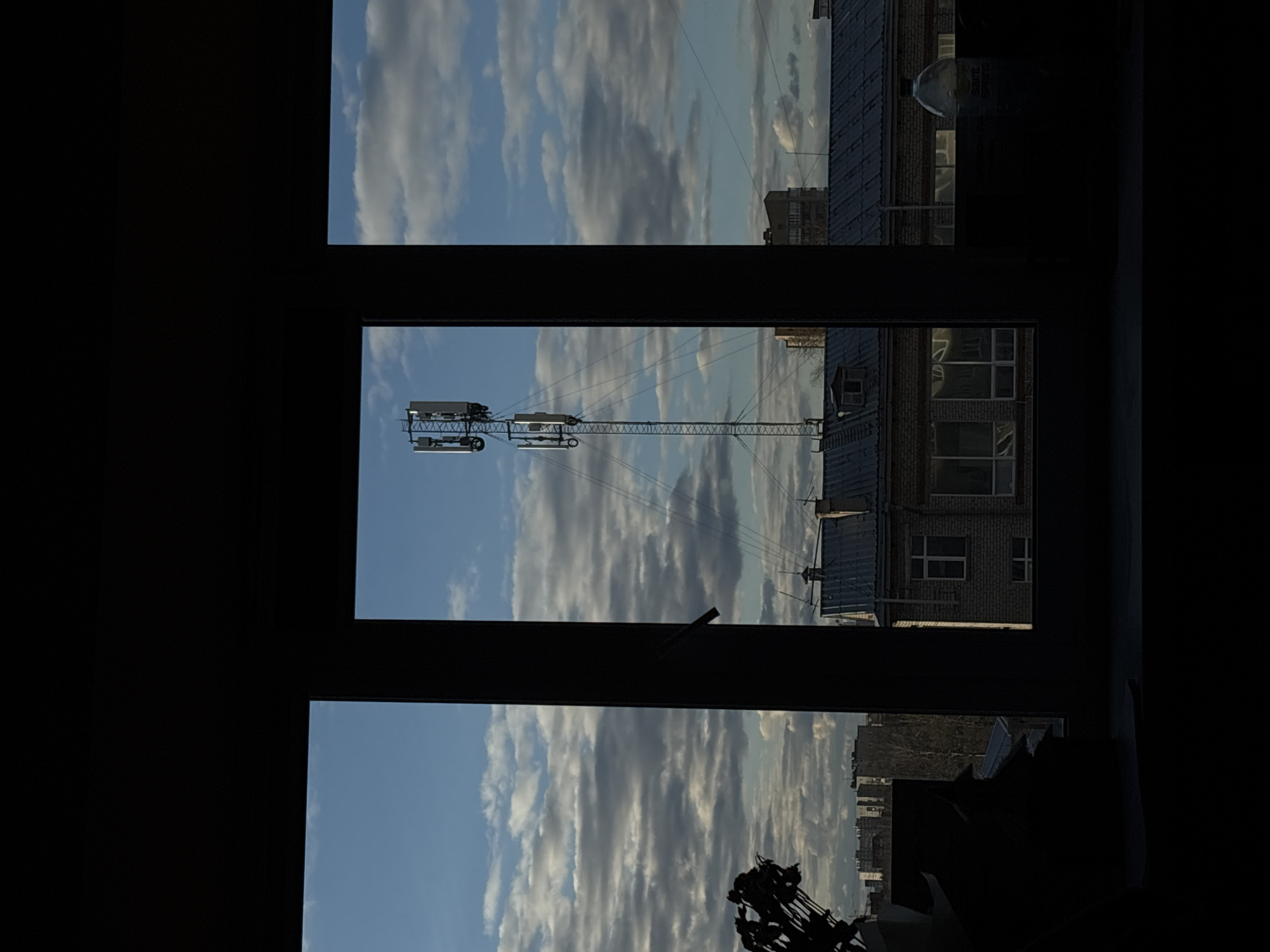 View from a window divided into three panes in Perm, Russia, shows a cloudy blue sky. A construction crane is visible through the central pane. Buildings are discernible through the window. The view is partially obscured by the frame of the window, which casts the sides of the image into shadow.