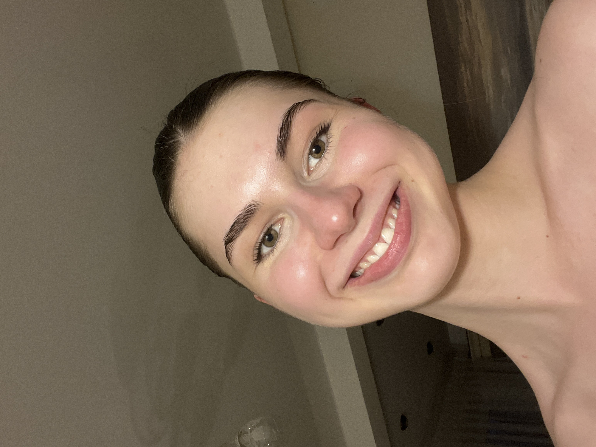 A close-up, angled shot features a fair-skinned woman with brown hair pulled back. She is smiling, revealing her teeth. Her eyebrows are dark and well-defined, and she has hazel eyes. Her skin appears clear and slightly flushed. The background consists of a beige wall and what appears to be a dark brown or black wooden surface. There is no indication of the location being Saint Petersburg, Russia.
