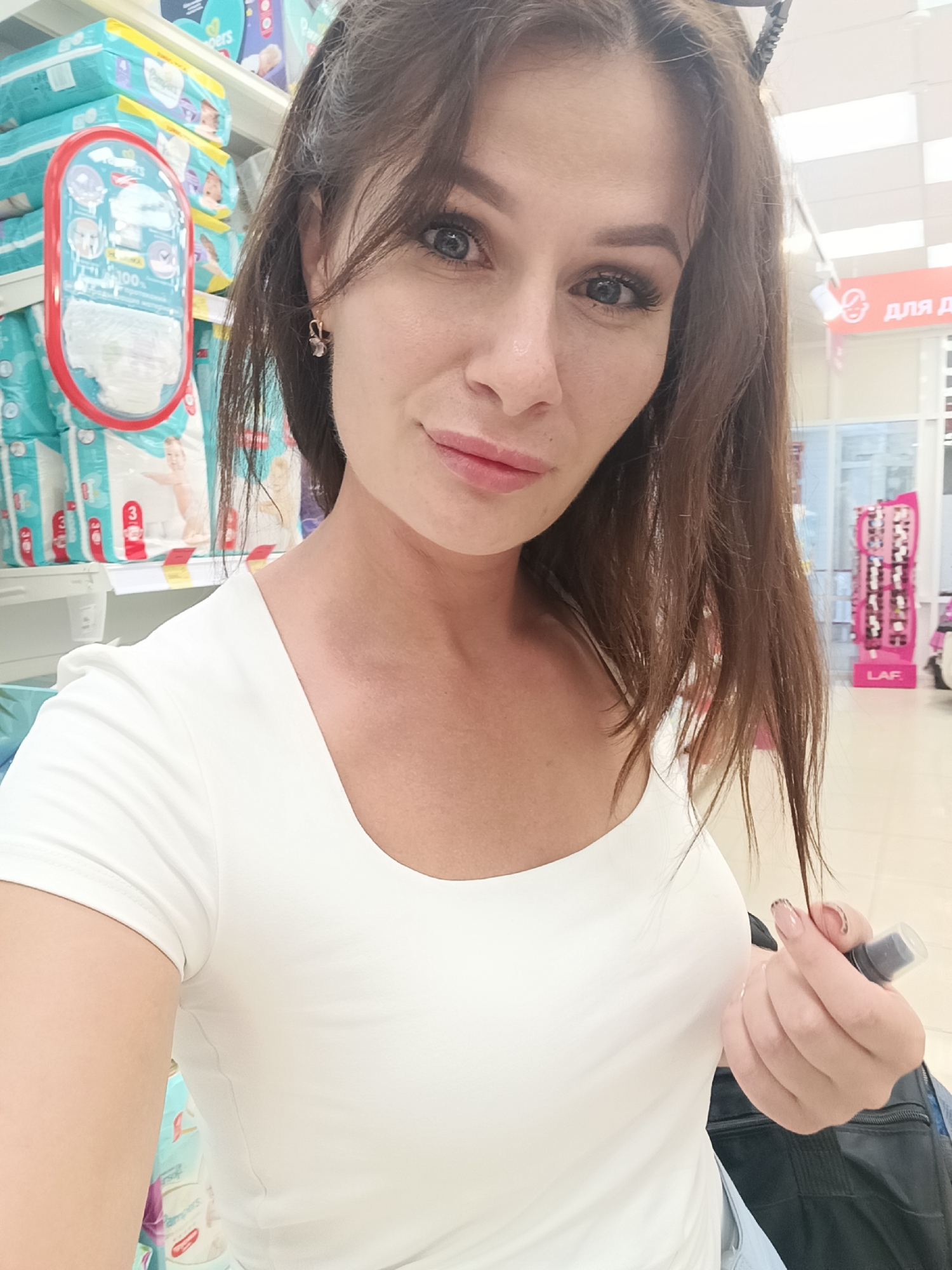 The image depicts a retail interior, likely a supermarket or baby goods store, located in the city of Gubkin, Russia. One adult female is positioned in the foreground, facing the camera and capturing a selfie. She possesses brown hair and blue eyes, and is wearing a white, short-sleeved top with a scoop neckline. Her right hand, exhibiting light-colored manicured fingernails with dark detailing, holds a small, dark-colored spray bottle. A dark bag strap is partially visible over her left shoulder. The background features extensive retail shelving units stocked with multiple packages of "Pampers" diapers, predominantly in turquoise packaging. A red-framed display containing a single diaper is visible on one shelf. Above the shelving, a red sign bears Cyrillic text, reading "ДЛЯ ДЕТЕЙ" (FOR CHILDREN). Further in the background, another red sign with "LAF" is partially visible, alongside a pink display rack containing numerous items. The ceiling consists of white panels with integrated artificial lighting, illuminating the scene brightly.