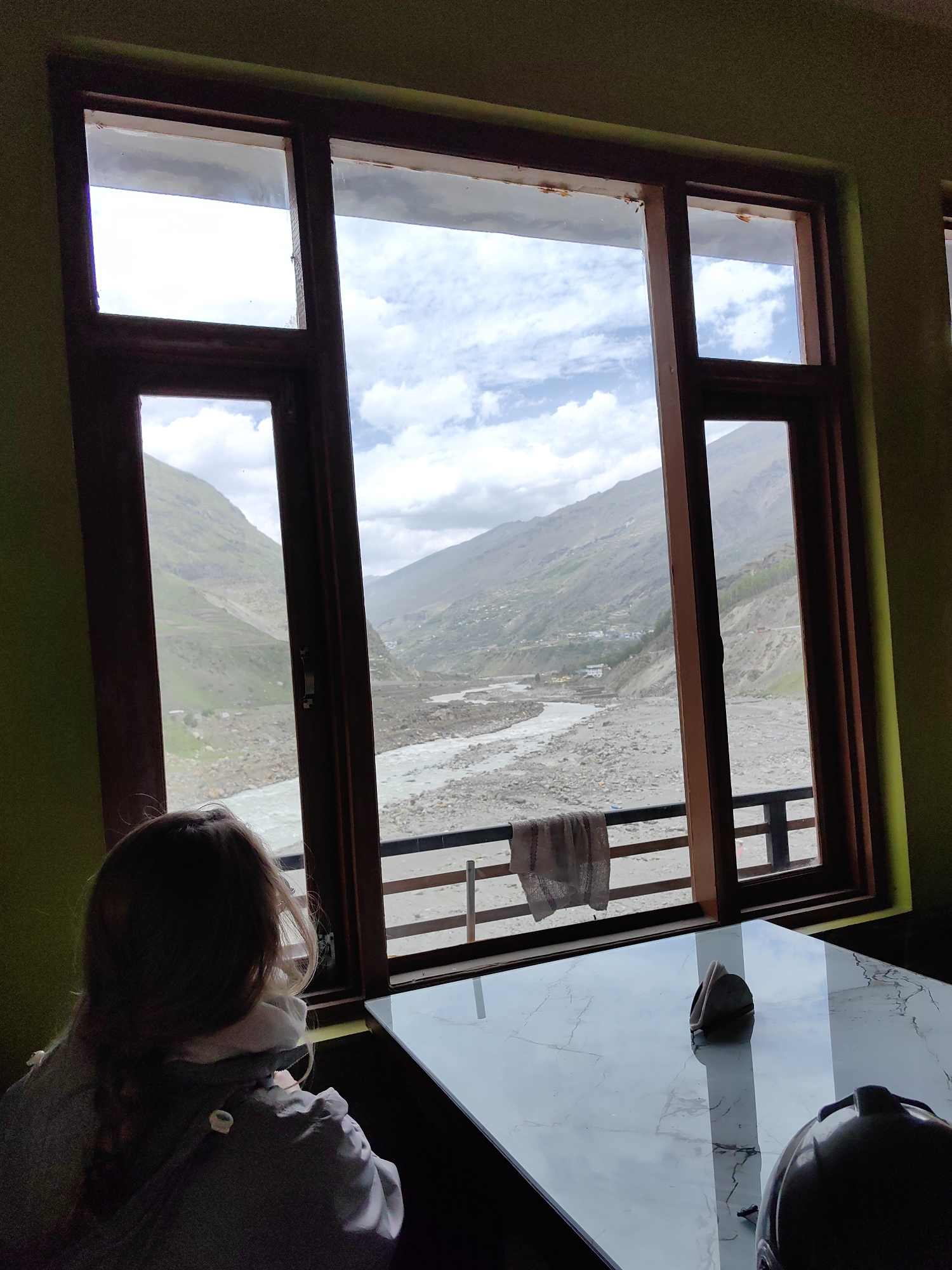 A person with light brown hair, viewed from behind, is sitting at a table in front of a large window. The window offers a view of a mountainous landscape with a river flowing through a valley. The person is wearing a light-colored jacket. On the table, a napkin holder is visible, as is a helmet in the foreground. The room walls are painted green. The exterior scene includes steep, vegetated slopes and a wide, rocky riverbed under a cloudy sky.