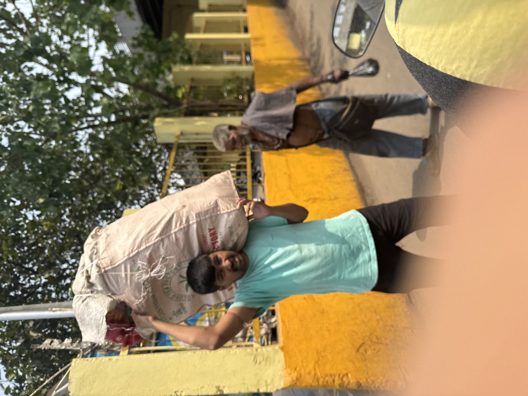 A young man carries a large sack on his shoulder in Kolkata, India. He wears a light blue t-shirt and black pants. An elderly man with bare torso and grey hair is walking alongside a building on the right. The background shows an urban environment with trees and buildings.