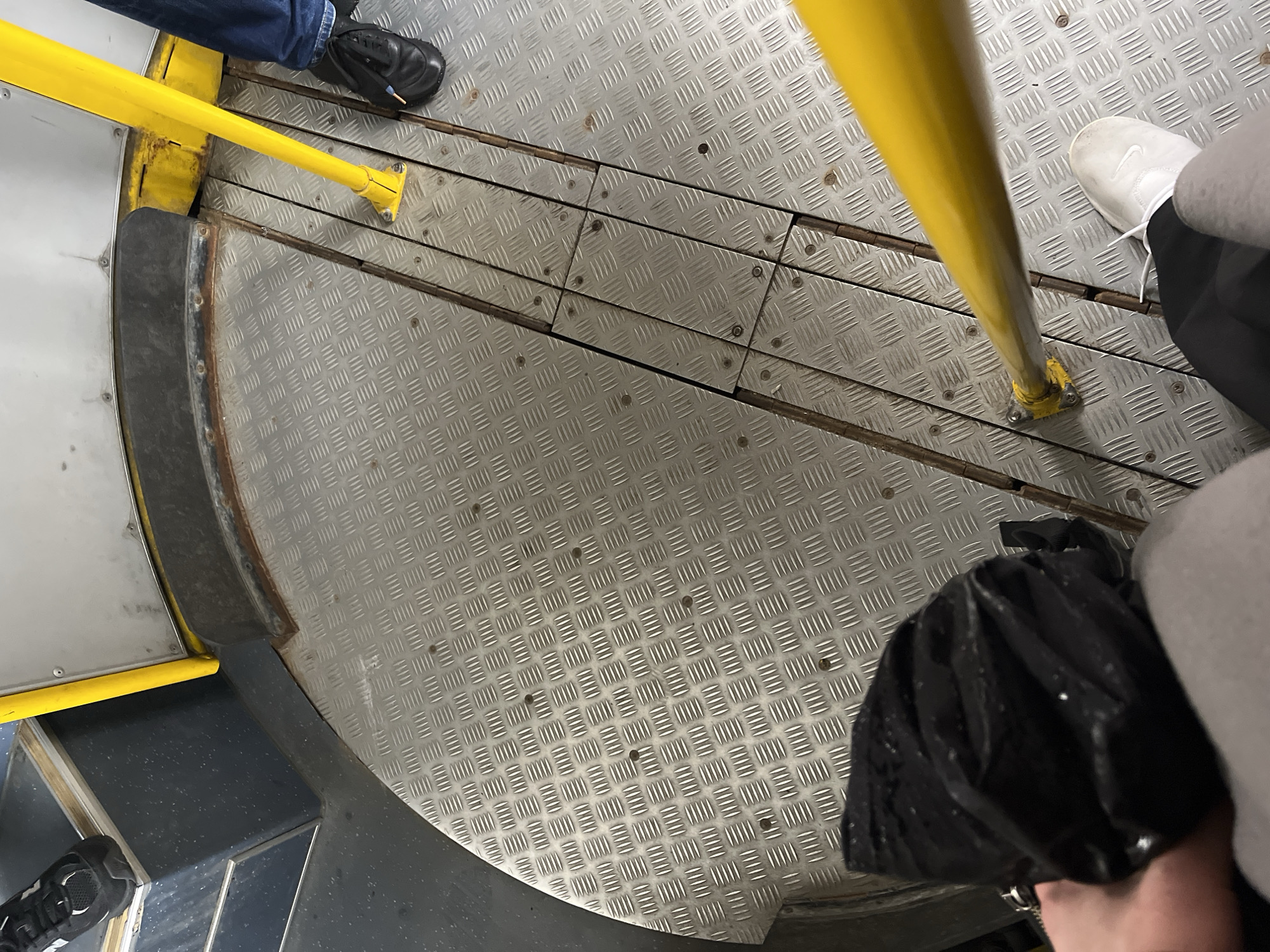 The image shows the interior of a public transport vehicle, possibly a bus or tram, with a focus on the floor and a yellow handrail. The floor is a textured metal plate with a diamond pattern, showing signs of wear and some rust. There are several visible screws holding the metal plates down. The area is partitioned by wooden planks beneath the metal plates in some sections. On the left, a black shoe with white accents is visible, partially on a dark blue textured surface. On the right, a person wearing blue jeans and a black shoe stands near a yellow handrail. Another yellow handrail is visible in the foreground, with a person's hand and a black, crinkled object (possibly a bag or coat) in the bottom left corner. A white shoe is also visible on the right side of the frame. The environment appears to be functional and utilitarian, common in urban public transport. There is no specific visual information in the image that would allow for a definitive determination of the location in the city of Minsk, Belarus.