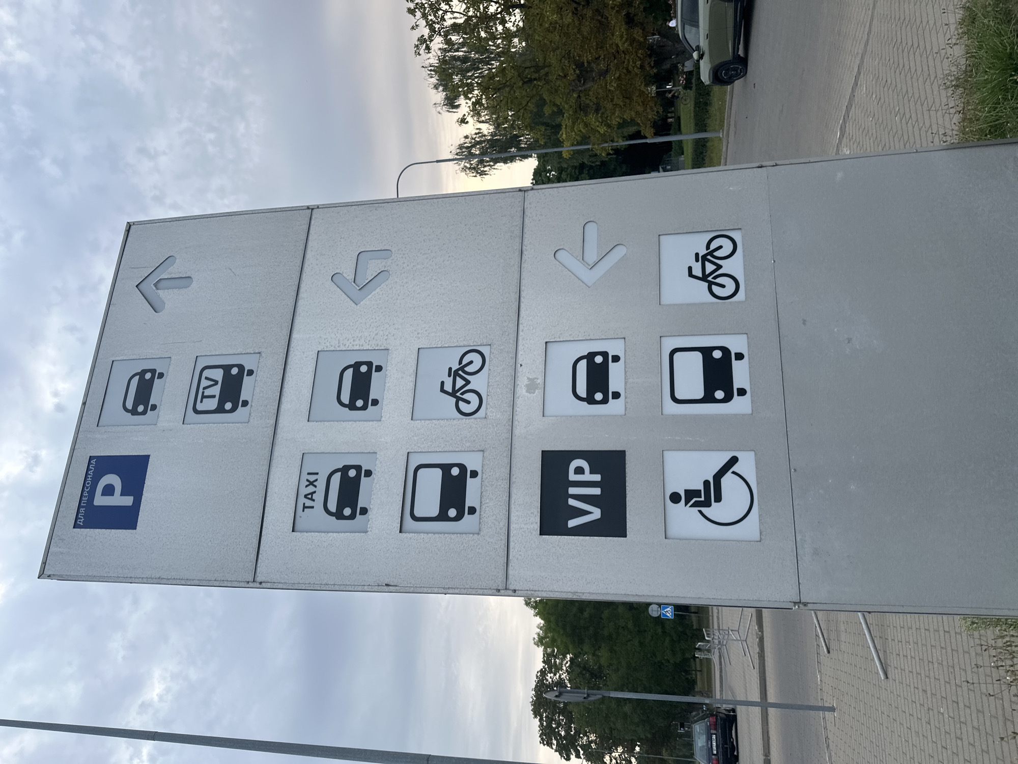The image shows a multi-panel signage structure located outdoors. The signage contains various pictograms and text indicating different parking areas and directions.
At the top, a blue square with a white "P" and the Russian text "ДЛЯ ПЕРСОНАЛА" (For Personnel) is visible, suggesting a parking area for staff. Below this, there are icons representing cars and a television, with an upward-pointing arrow to the right of the car icon.
Further down, a section labeled "TAXI" with a car icon is present. To its right, another car icon and an arrow pointing left are displayed. Below the taxi section, there's an icon of a bus, and to its right, a bicycle icon.
Another section prominently displays "VIP" in large white letters within a black square, indicating a VIP area. To the right of the VIP section, there's a car icon and a left-pointing arrow. Below the VIP section, an icon representing a person in a wheelchair is shown, and to its right, a bus icon and a bicycle icon are present.
The signage is made of metal and appears to be weathered. The background includes a cloudy sky, trees, a street with cars, and a paved pedestrian area. A street lamp is visible on the right side of the image.
The context provided indicates this location is in Minsk, Belarus. Without more specific visual cues, it is not possible to determine the exact location within Minsk, but the signage suggests a public transportation hub, parking facility, or a large complex with designated areas. No people are clearly visible in the immediate vicinity of the signage, though a dark-colored car is parked in the background on the left, and a green SUV is visible on the road to the right. A pedestrian crossing sign is visible on the left side of the image.