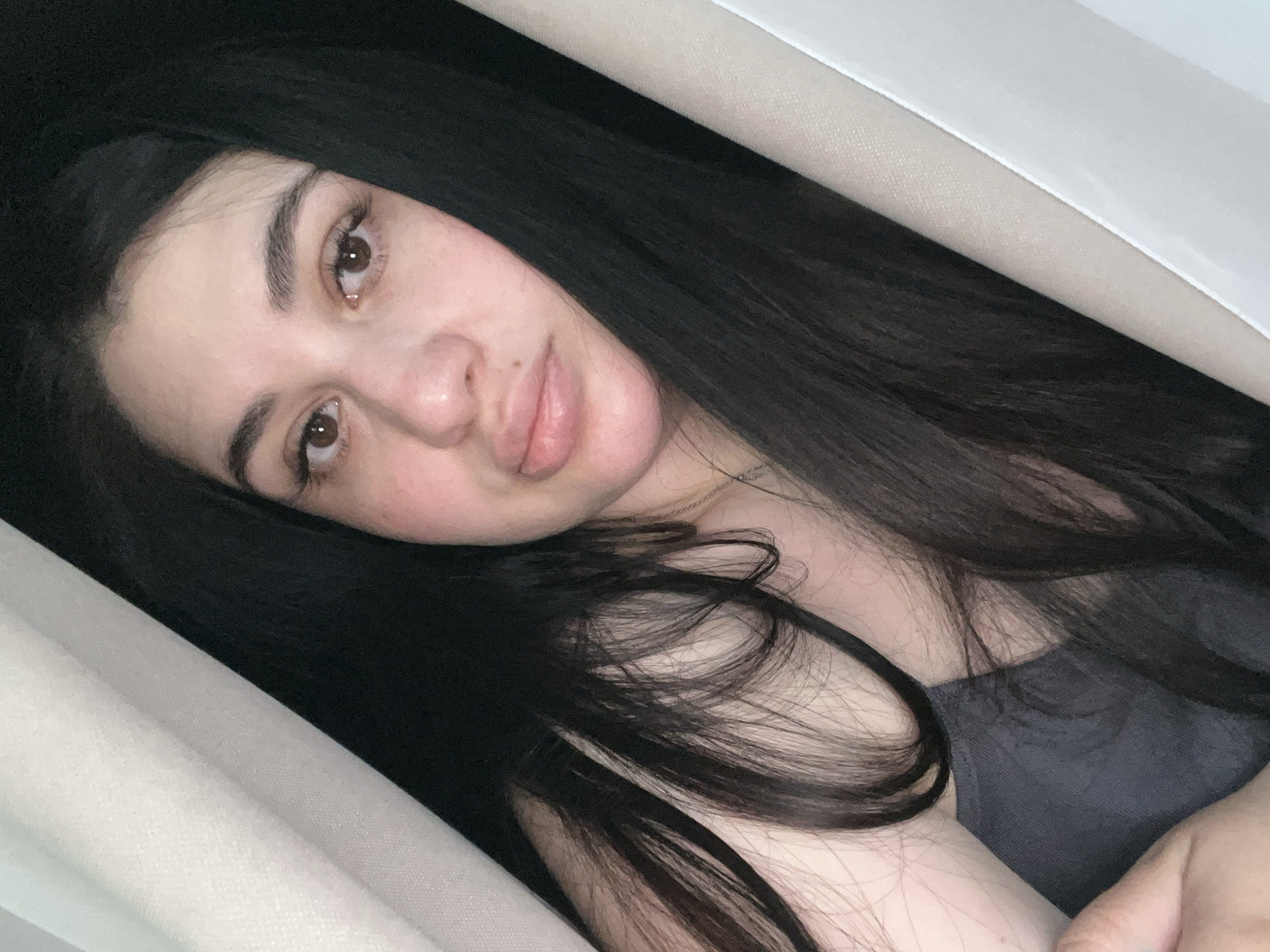 A young woman with long, dark hair is lying down, likely indoors, based on the presence of what appears to be white fabric or upholstery surrounding her. Her face is partially angled towards the camera, showing her brown eyes, light skin, and slightly parted lips. She is wearing a gray top. The image does not provide sufficient detail to confirm her location, and specifically, cannot verify her presence in the city of Chaikovsky, Russia. No actions, interactions, or distinct objects can be determined from the image.