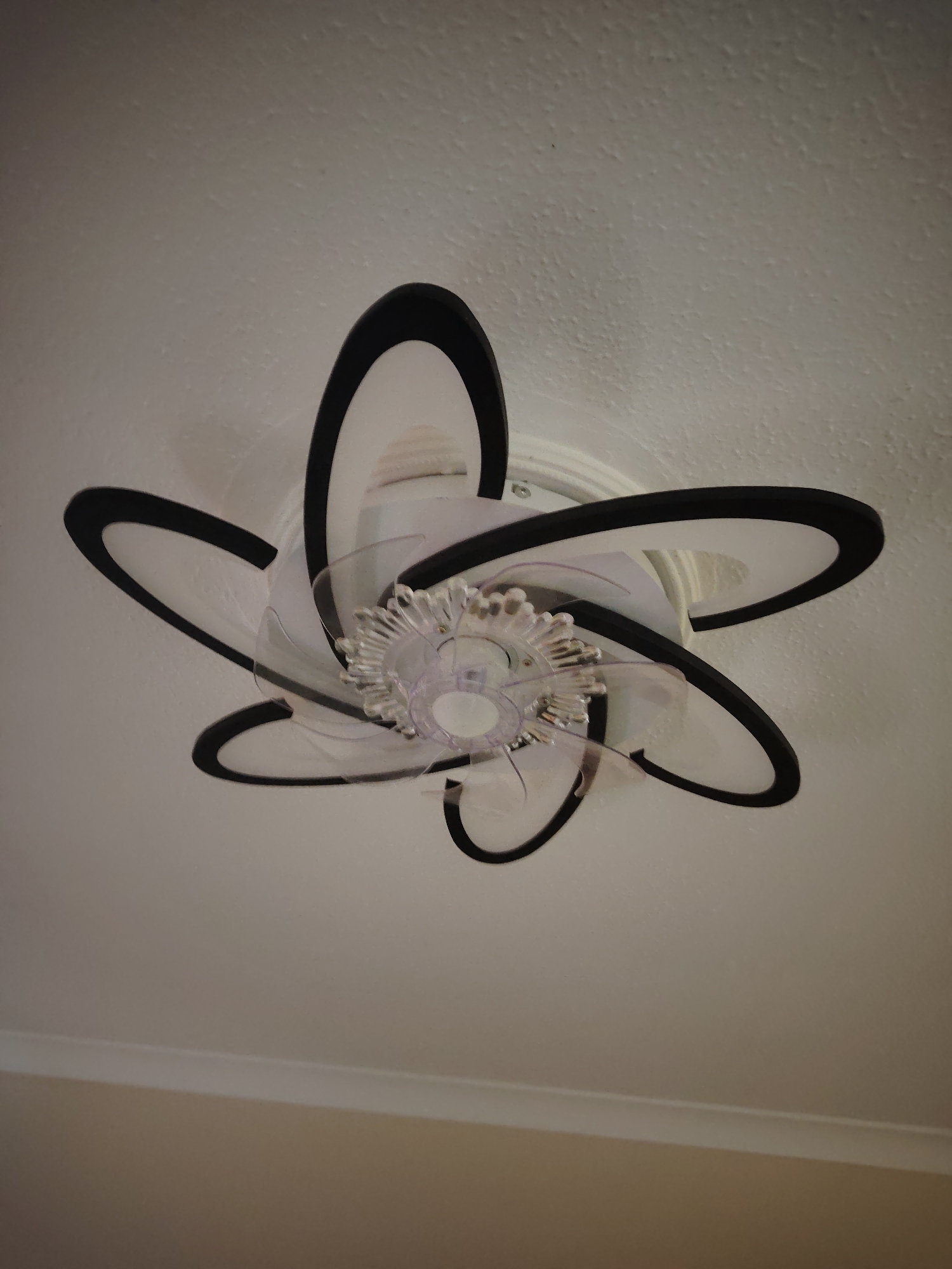 An overhead view captures a contemporary ceiling fixture mounted flush against a textured, light-colored ceiling. The fixture, observed in Huntingdonshire, United Kingdom, functions as both an illumination source and an air circulation device. It comprises a central fan unit equipped with multiple clear, curved plastic blades. Surrounding the fan's hub is a decorative, multi-faceted clear plastic or crystal-like element. Six large, curved, petal-like components are arranged radially around this central assembly. Each component features a distinct black outer trim and a white translucent surface, indicative of embedded LED light panels. A segment of a light beige wall is visible in the lower portion of the image, separated from the ceiling by a white molding strip.