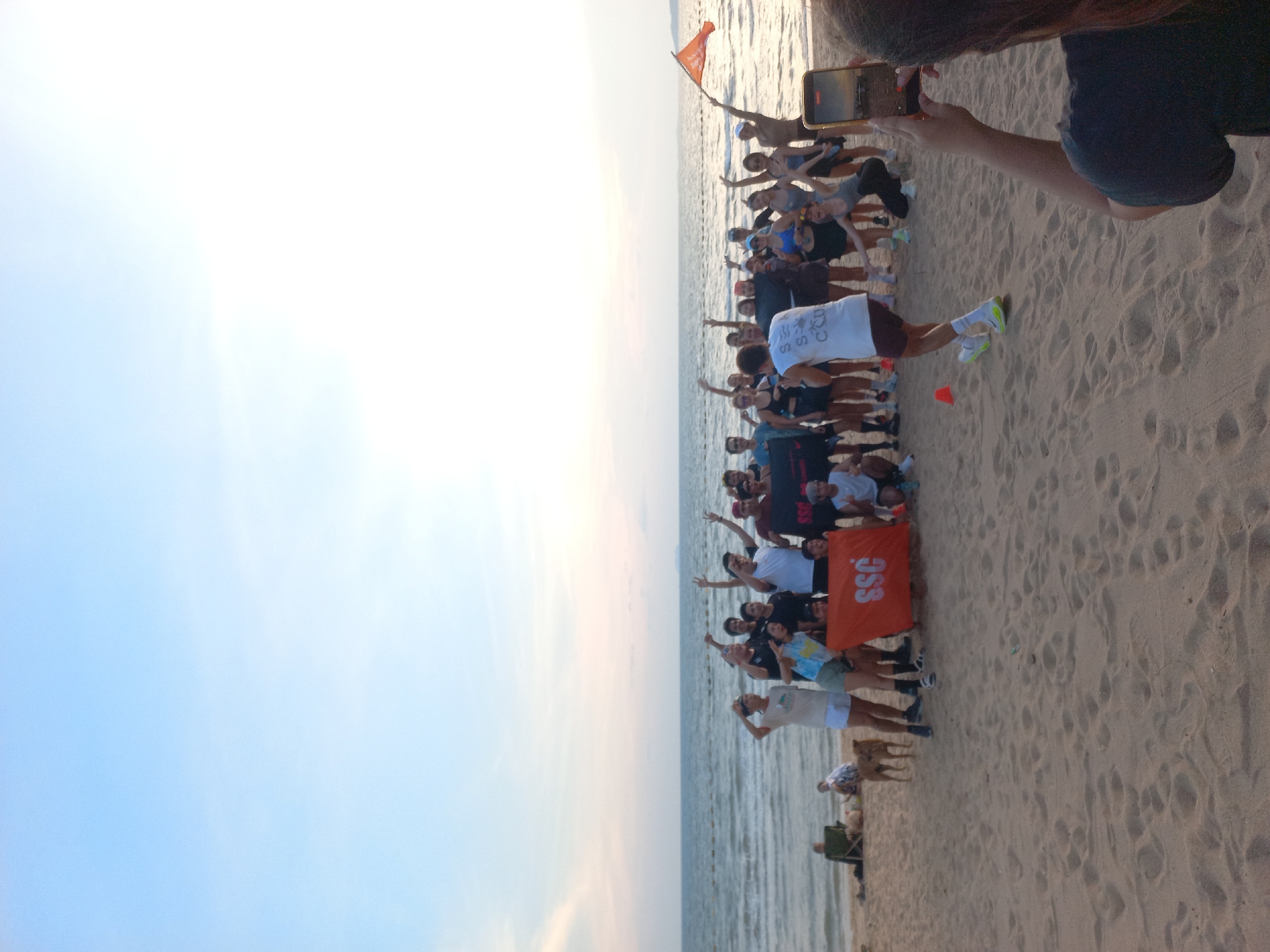 A large group of people are gathered on a sandy beach with a body of water in the background, likely the sea. The scene appears to be at Pattaya City, Thailand. The sky is partially visible with light tones. A person is taking a video or picture with their phone of the group. The group includes individuals in various attire, with some holding an orange flag bearing the letters "SSC". Some members of the group have their arms raised in the air. The beach is visible, exhibiting footprints.