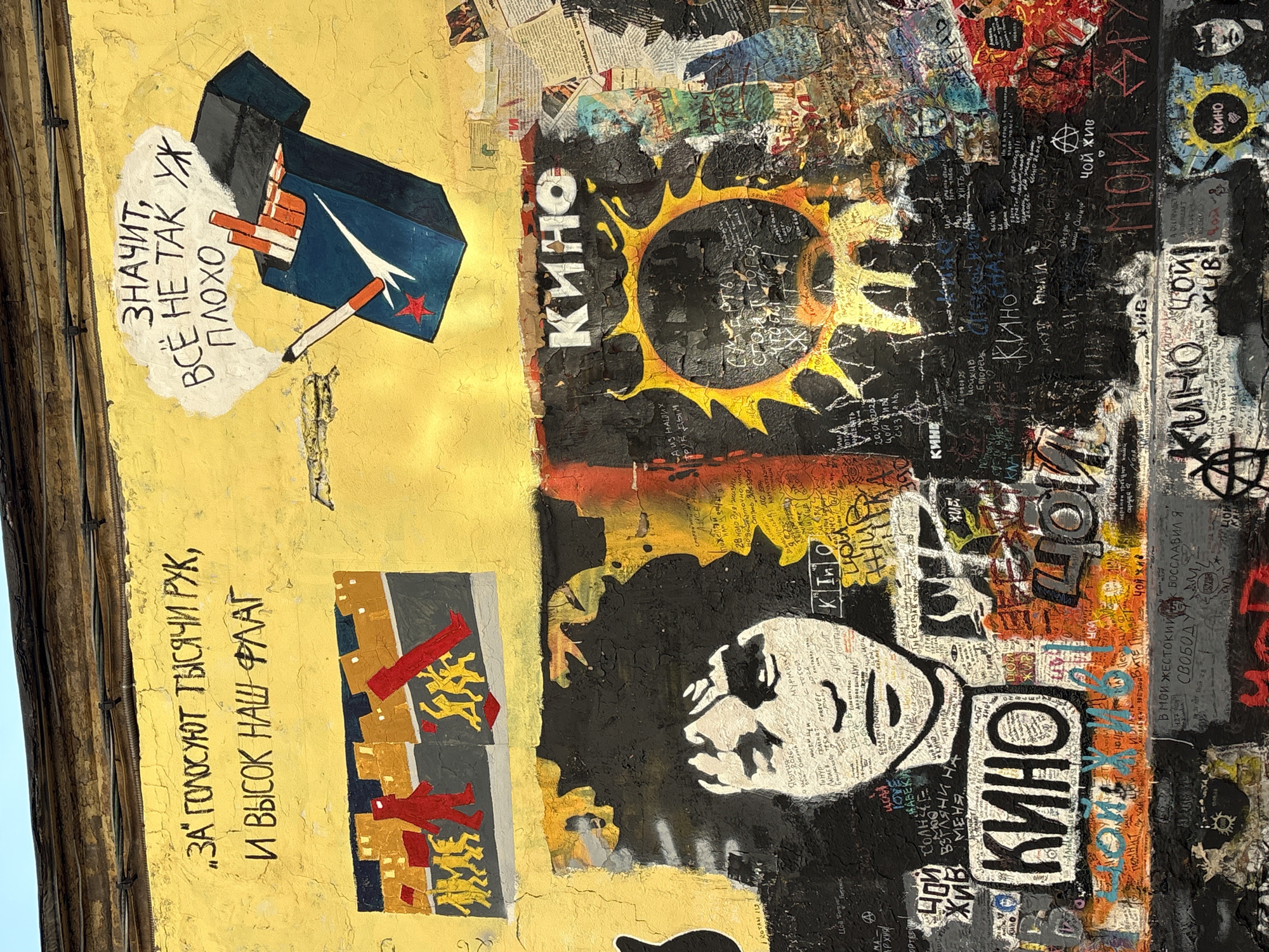The image is a close-up of a vibrant and weathered mural painted on a yellow stucco wall in Moscow, Russia. The mural depicts stylized figures, text in Russian, and a portrait of a person with dark, curly hair.
In the upper left, black Russian text reads: "ЗА ГОЛОСУЮТ ТЫСЯЧИ РУК, И ВЫСОК НАШ ФЛАГ." Below this, a graphic shows a line of figures walking towards buildings.
To the right, a speech bubble with Russian text "ЗНАЧИТ, ВСЁ НЕ ТАК УЖ ПЛОХО" (Meaning, not all is so bad) floats above a drawing of an open pack of cigarettes and a lit cigarette.
The central and lower portions of the mural feature a large, stylized black and white portrait of a person's face. The word "КИНО" (Kino) appears multiple times in prominent white lettering. A large, jagged yellow and black sun-like image is also depicted. The wall is covered in layers of paint, graffiti, and possibly pasted paper, indicating a history of additions and alterations.
This specific mural is located at the "Wall of Viktor Tsoi" (Стена Цоя) on Arbat Street in Moscow. Viktor Tsoi was a Soviet rock musician and leader of the band Kino, and this wall has become a memorial site for him.