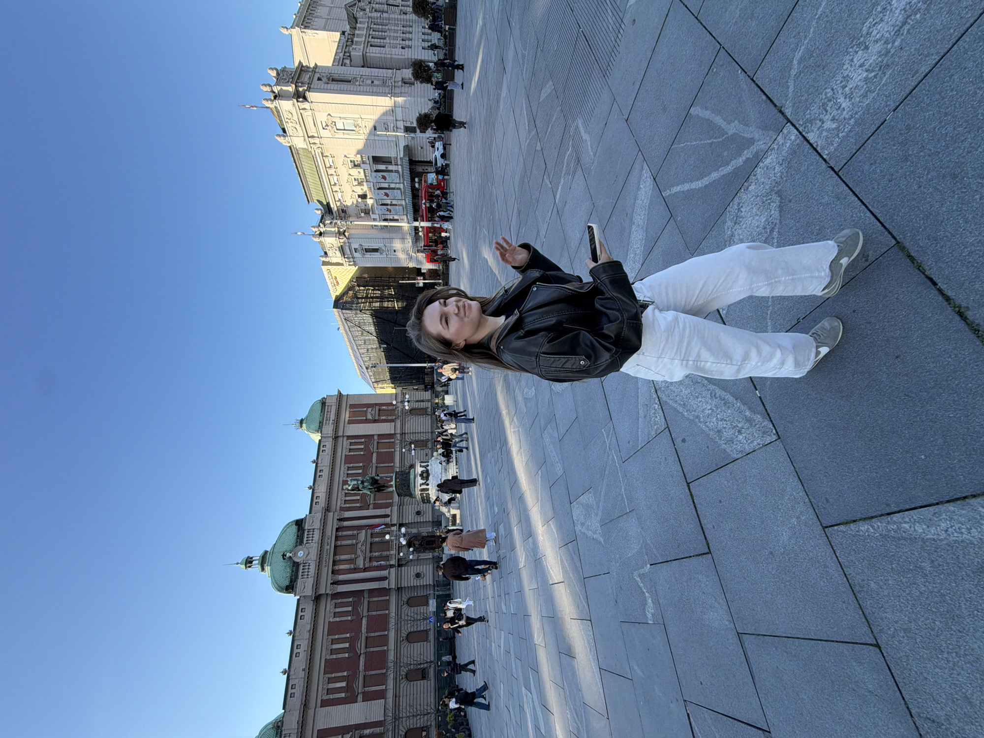 A young woman stands on a paved square in Belgrade, Serbia, holding a smartphone in her right hand, in an area filled with pedestrians. She is wearing a black leather jacket, white pants, and grey sneakers. In the background, two buildings can be identified: the National Museum of Serbia on the bottom, and the National Theatre on the top. The sky is clear and blue. The pavement consists of large, grey tiles. The image is rotated counter-clockwise.