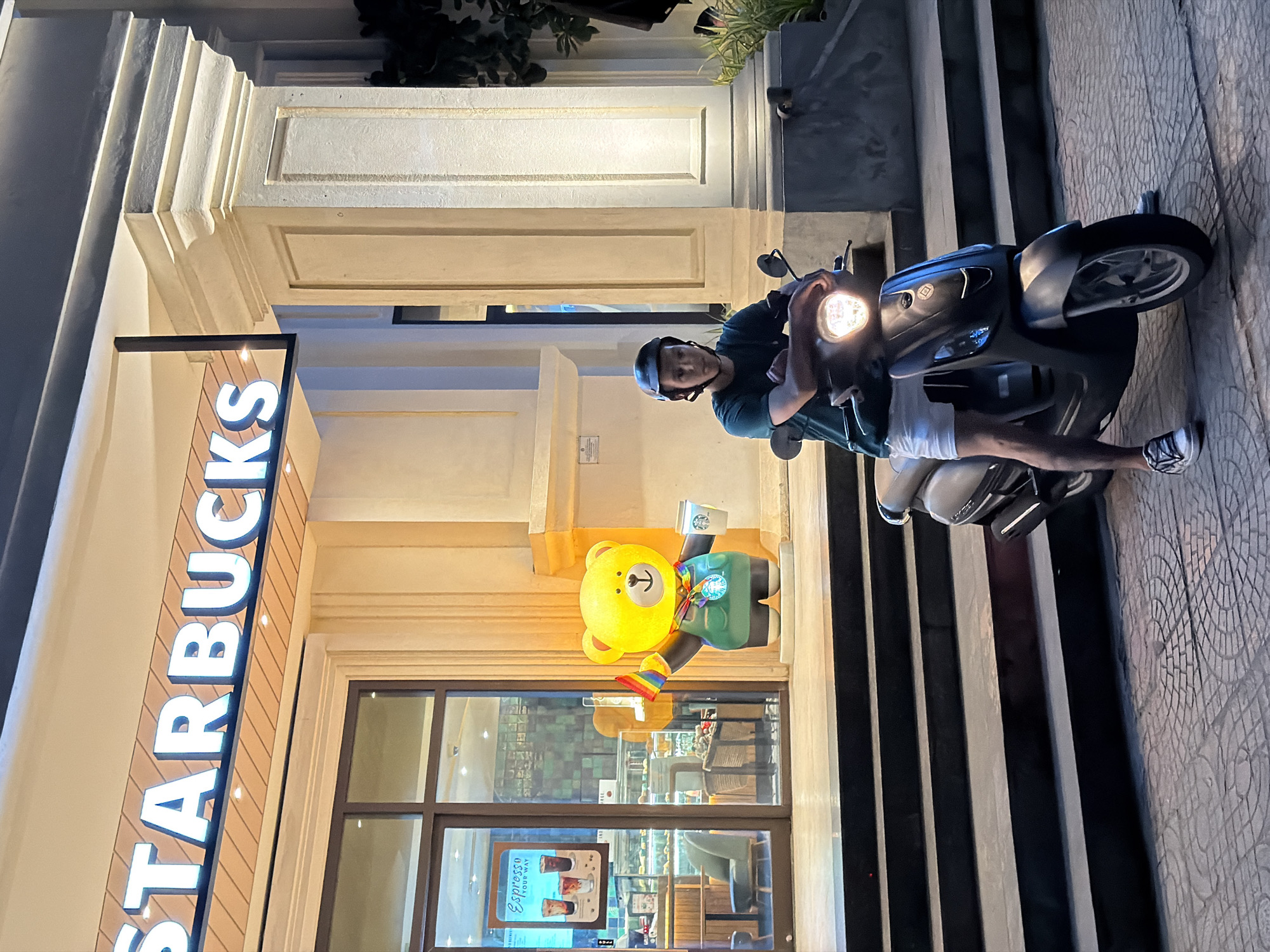 A person is sitting on a scooter in front of a Starbucks in Da Nang, Vietnam. The Starbucks sign is lit up and visible above the entrance. To the left of the entrance, a large yellow bear statue is standing, holding a rainbow flag and a cup. The person on the scooter is wearing a green t-shirt, white shorts, a helmet, and sneakers. They are looking towards the camera. The scooter's headlight is on. The scene appears to be at night, with ambient lighting illuminating the storefront and the scooter. The ground is paved with interlocking stones. The facade of the building has decorative columns.
