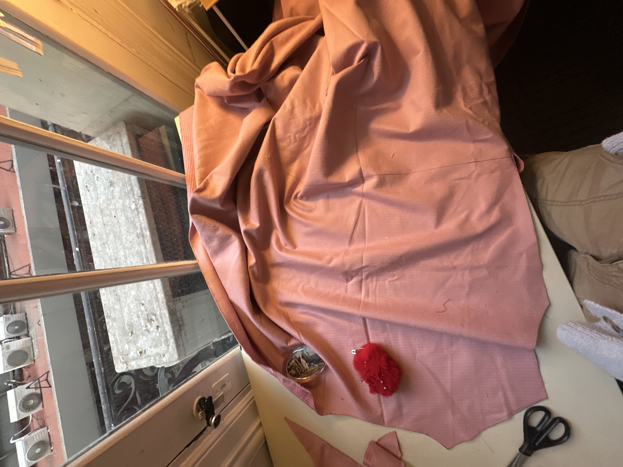 The scene is indoors, presumably a workshop or home interior, adjacent to a window. The window offers a view of an exterior, which includes elements of a building's facade, specifically multiple air conditioning units mounted on a building in the city of Buenos Aires, Argentina, as well as a metal balcony.
Inside, the predominant object is a large piece of pink fabric, draped across the surface. Visible on the surface with the fabric are a small, clear container, a pincushion of red color with pins, and a pair of scissors. The fabric appears to be in the process of being cut, as indicated by the presence of the scissors and smaller cut pieces of fabric. The environment is lit by daylight coming through the window.