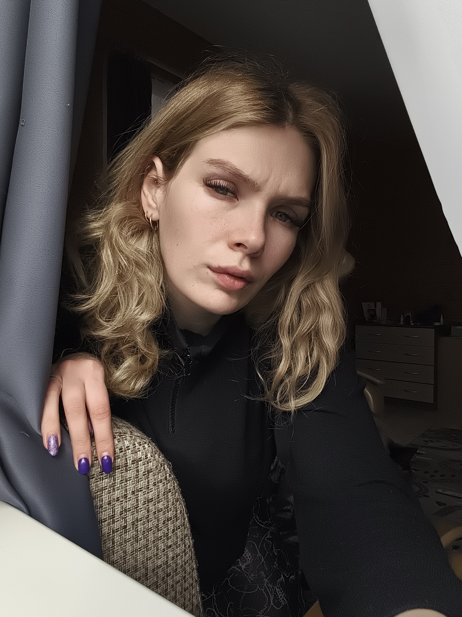The image depicts a young woman with light blonde, wavy hair, posing indoors near a window covered by a gray curtain. She is wearing a black long-sleeved shirt with a high collar. Her nails are painted in purple hues, with some featuring glitter. The woman has a fair complexion and appears to be looking directly at the camera with a slight frown. The background includes an attic-style ceiling and a dresser with drawers in the dimly lit room. While the prompt requests the location in Русский Вожой, Russia, the image itself doesn't provide any evidence to definitively confirm this location.