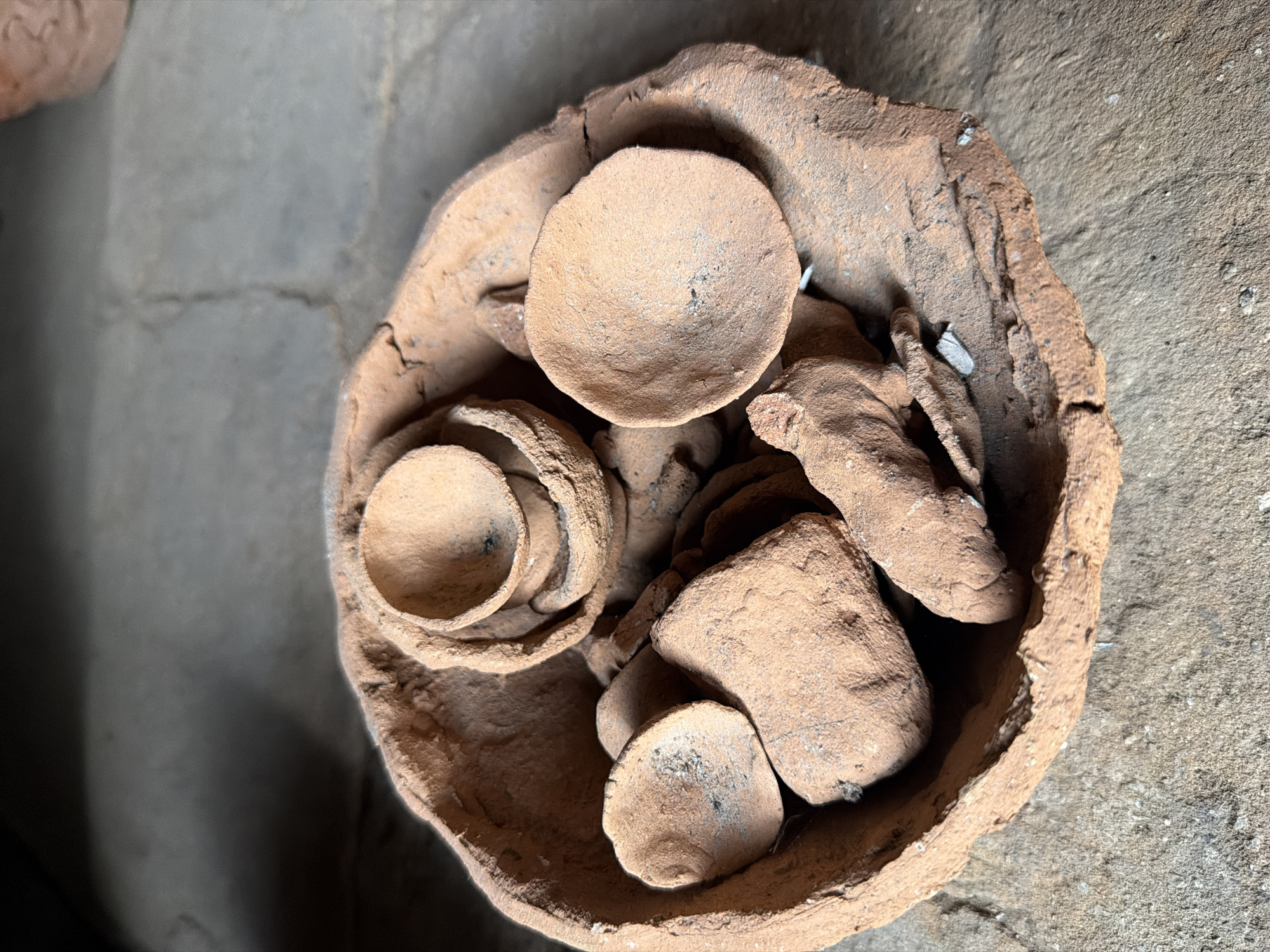The image shows a top-down view of a rough, hand-made pottery bowl filled with smaller, similarly crafted clay items, likely cups or small bowls. The pottery is a reddish-brown color, and appears unglazed with a coarse texture. The bowl sits on a rough, concrete-like surface. The image was taken in Makatubani, Georgia.