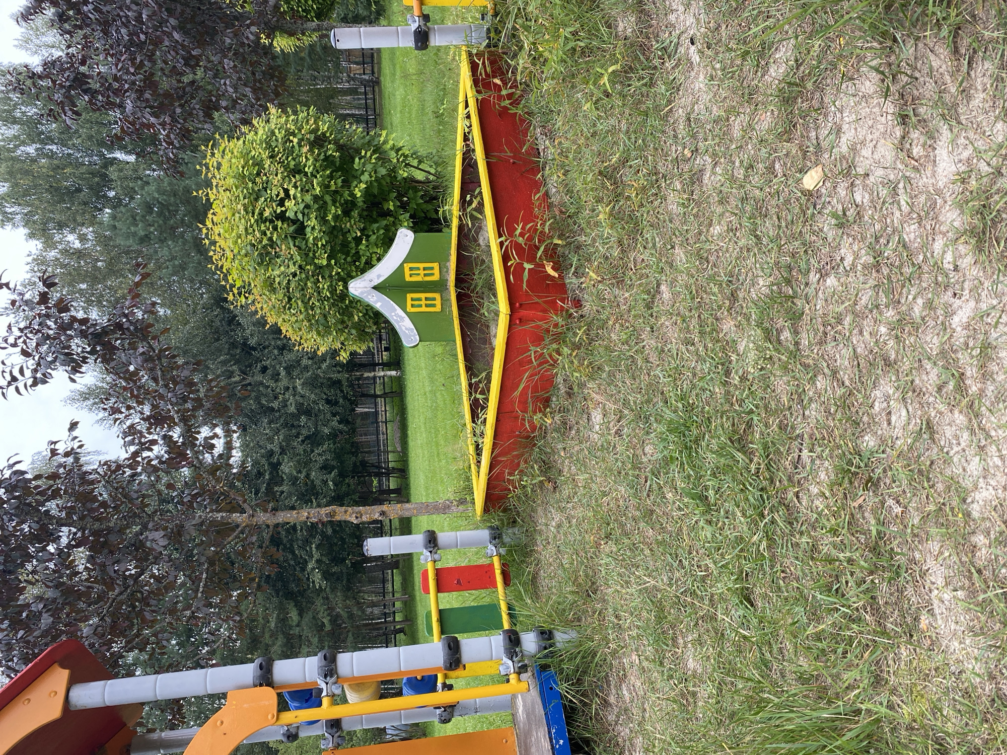 An outdoor scene in Chulis, Russia, features a playground area with a sandbox and a colorful play structure. The sandbox, rectangular and red with a yellow border, contains sand and some overgrown weeds. Adjacent to the sandbox is a small, green, house-shaped structure with white trim and two yellow windows. A part of a multi-colored playground structure, consisting of yellow and orange plastic elements and gray metal poles, is visible on the left side of the image. Several trees with dark red and green foliage and a dense green bush are present in the background. The ground is covered with grass and sand. The overall environment suggests a public park or a recreational area.
