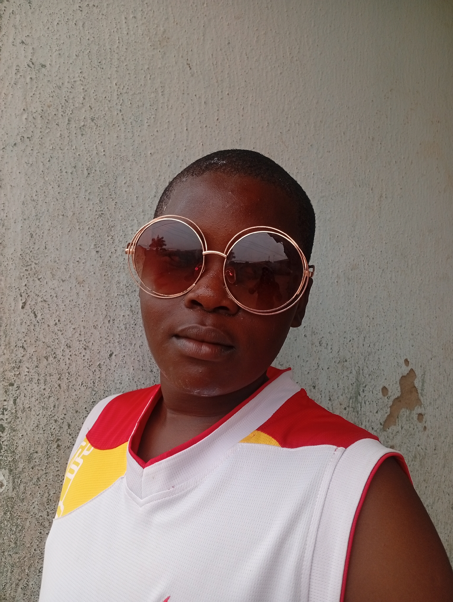 A dark-skinned person with a shaved head is wearing large round sunglasses with gold frames, a white sleeveless athletic shirt with red and yellow accents, and standing in front of a light gray textured wall. The reflection in the sunglasses suggests an outdoor setting with trees. The location is Sabon Tasha, Nigeria.