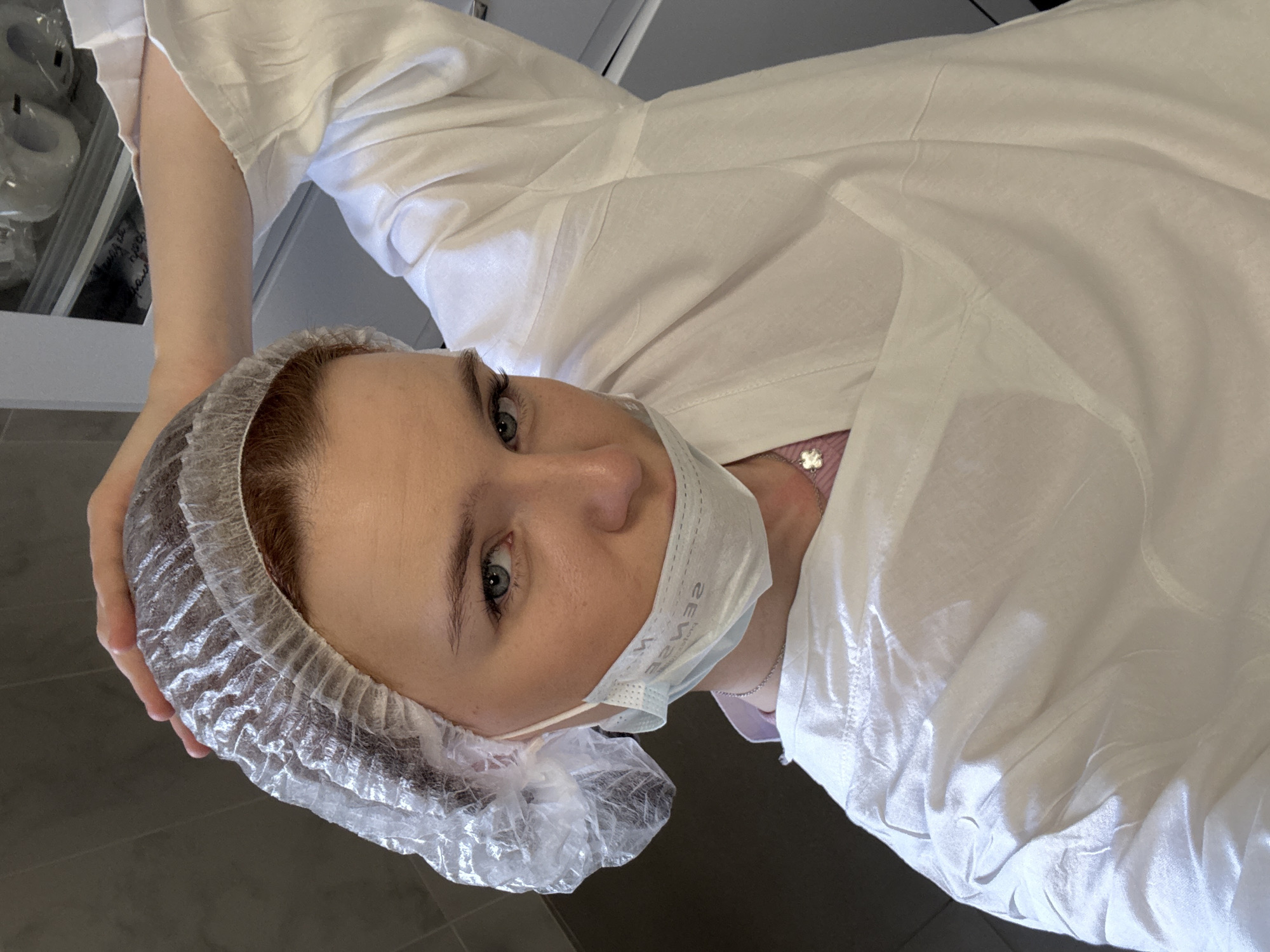 A woman with fair skin and light eyes, wearing a white disposable surgical cap and a light blue surgical mask, is positioned facing the camera. She is wearing a white lab coat, and a glimpse of a pink garment is visible beneath the coat near her neckline, along with a silver chain. Her left hand rests on her head, next to the cap. The background includes what appears to be a cabinet, containing items. The flooring visible in the scene is dark and smooth. The location is not identifiable as Saint Petersburg, Russia.