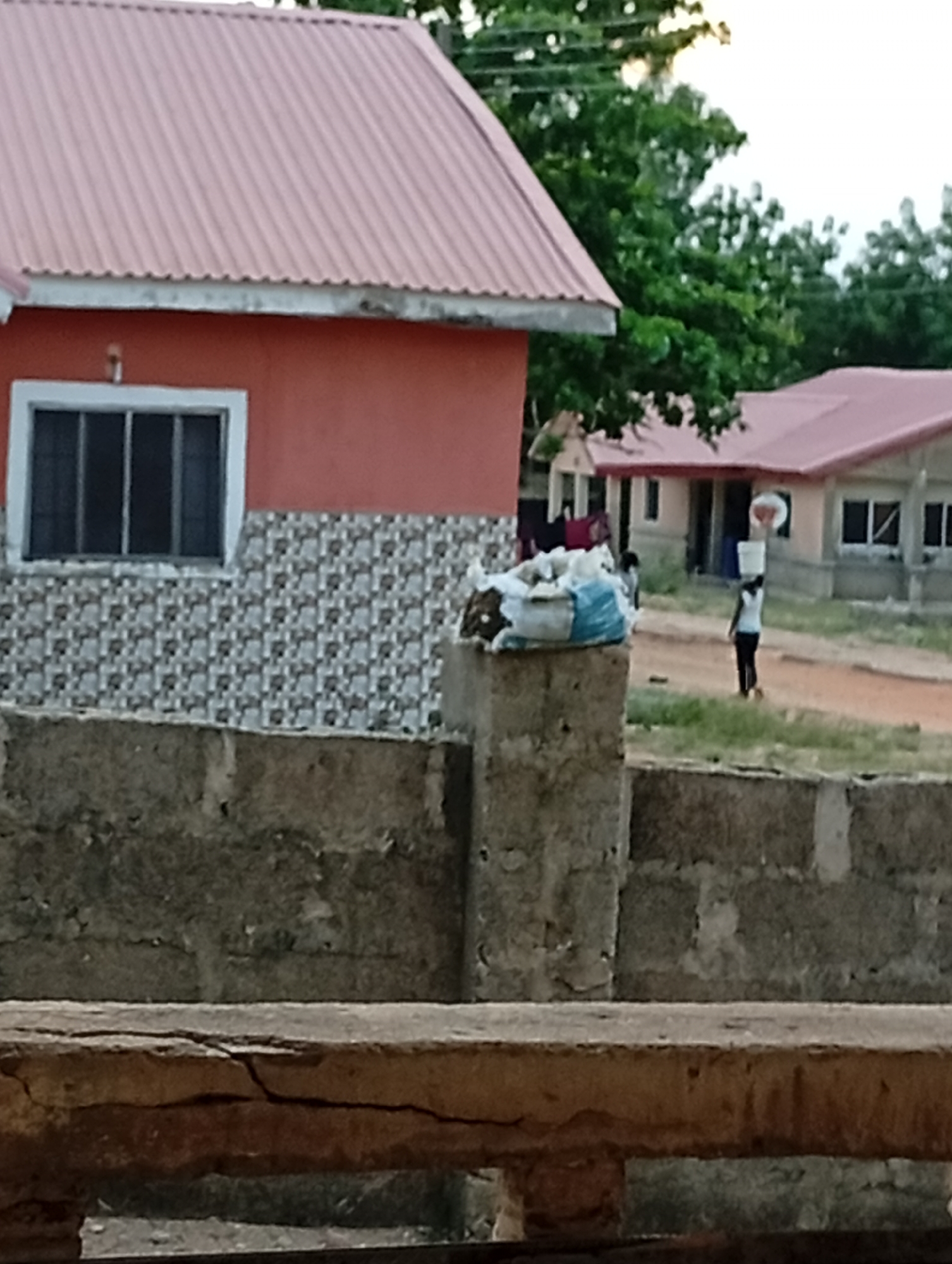 The image captures a scene in Adankolo, Nigeria, featuring a red-painted building with a pink corrugated metal roof and a tiled facade beneath a window with white trim. A concrete block wall is in the foreground, topped by a block post supporting what appears to be bags. In the background, a woman is carrying an item on her head, walking on a dirt path towards another red-roofed building. Trees are visible in the background.