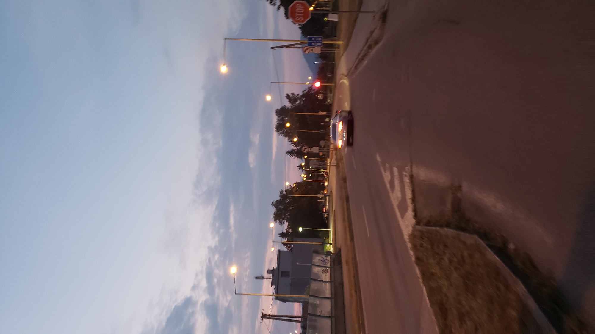 The image depicts a street scene in Prešov, Slovakia, during twilight. A silver car is driving down the road, with its headlights on. Streetlights illuminate the scene, casting a warm glow on the asphalt. There are buildings and trees on either side of the road, and a stop sign is visible on the right side of the image. The sky is a dusky blue with scattered clouds.