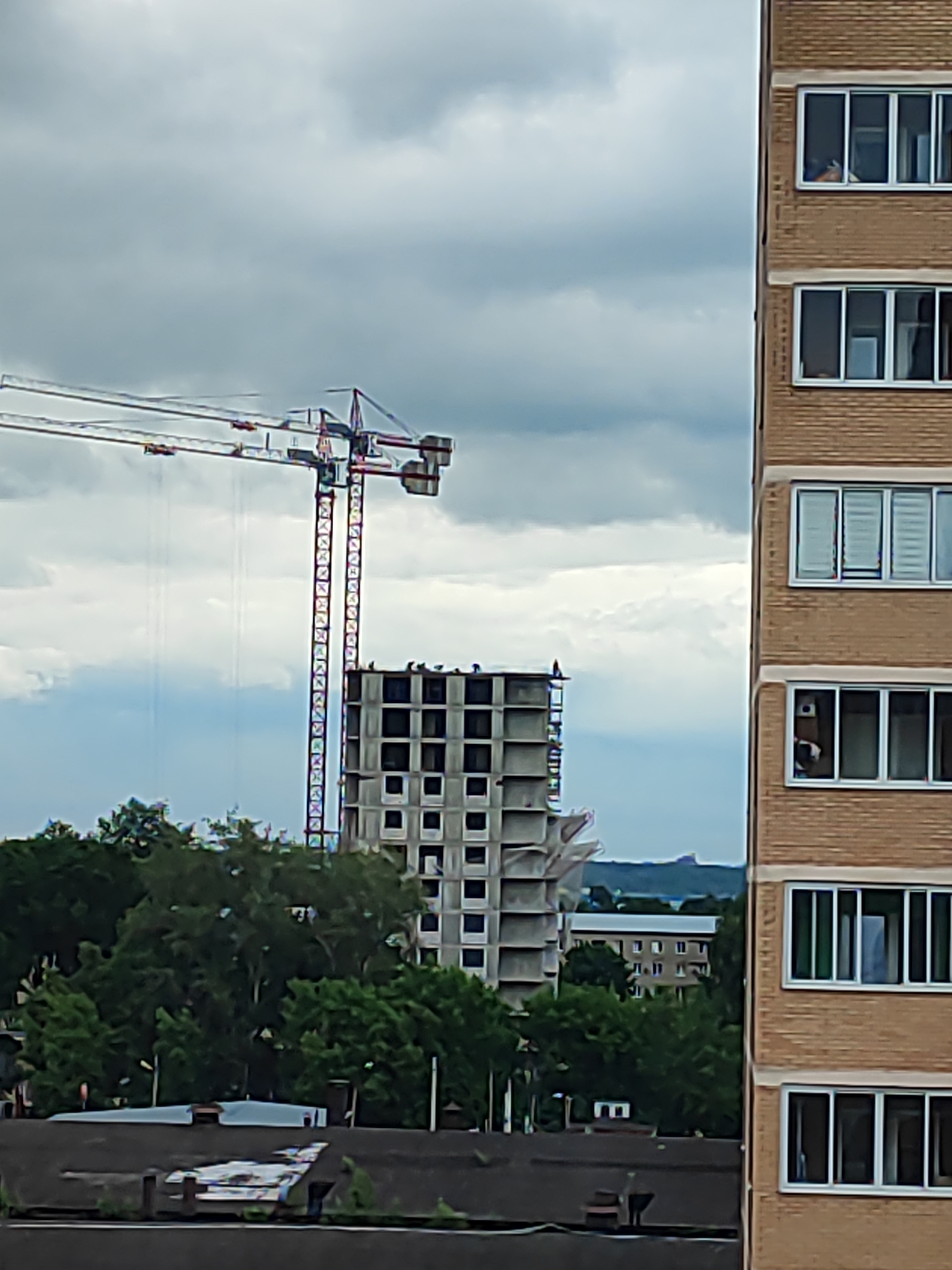 The image shows a construction site with a tall crane and an unfinished building. To the right, there is a brick building with several floors and windows. The sky is cloudy. The image was taken in the city of Shchyolkovo, Russia, on a cloudy day. The scene depicts urban development and construction activities.