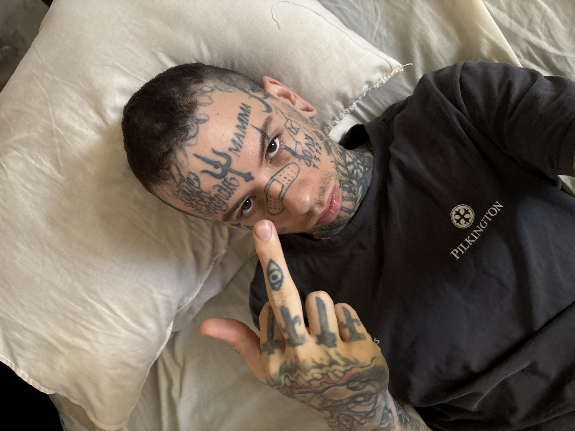 This medium shot captures a young individual lying on a light-colored pillow, looking directly at the viewer. The person is heavily tattooed, with extensive ink covering their face, neck, and hands. Prominent face tattoos include the word "MAMMA" on the forehead, a trident-like symbol near the left eye, various scripts, and other geometric designs. A flesh-toned band-aid is affixed to the bridge of their nose.
The person is wearing a dark grey or black t-shirt, on which a white logo and the word "PILKINGTON" are visible on the left chest area. Their right hand is raised and oriented towards the camera, with the middle finger extended in a gesture of defiance or casual disrespect. This raised finger also displays an eye symbol tattoo, while other fingers have distinct sword-like designs and the partial word "MAS" visible on the knuckles. The setting appears to be an indoor environment, possibly a bedroom or living space, indicated by the pillow. The lighting suggests daytime, though no specific light source is visible. While the image itself provides no explicit location cues, the context states this media file is from Buenos Aires, Argentina. The overall mood conveyed by the pose and gesture is one of bold nonchalance.