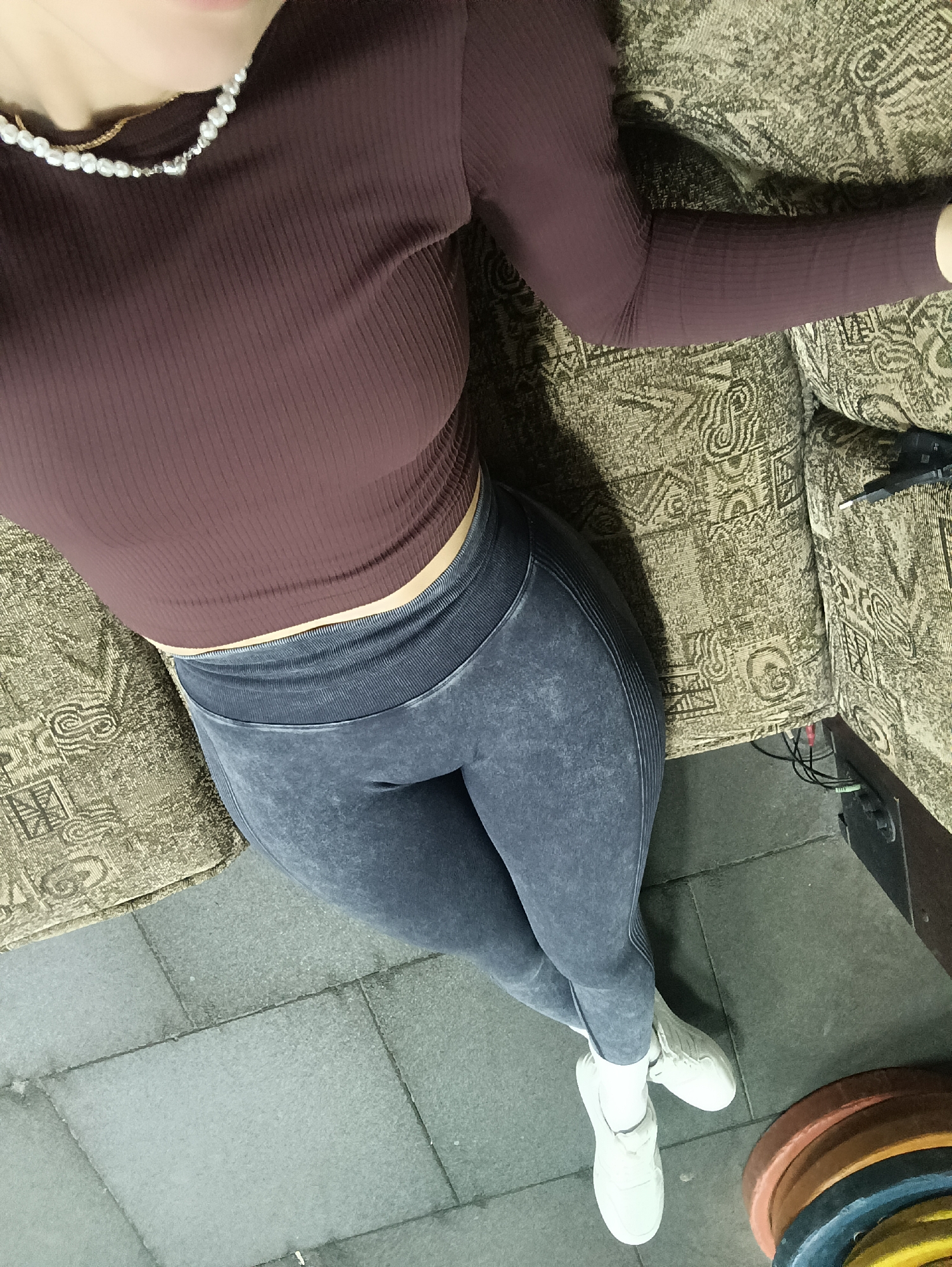 The image shows a person lying on a patterned couch. They are wearing a long-sleeved, ribbed brown top, gray leggings, and white sneakers. The person is also wearing a pearl necklace. The setting appears to be a gym, as weight plates are visible on the floor next to the couch. The floor is tiled.