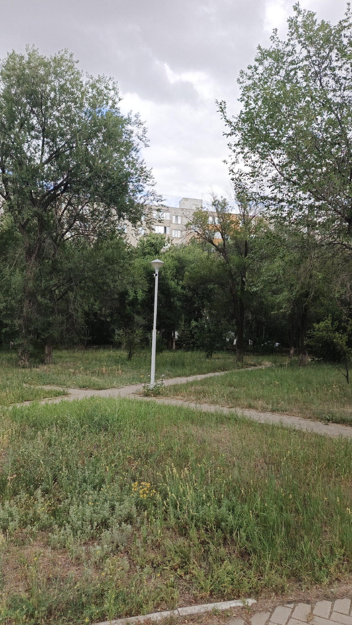 The image depicts a paved pathway intersecting a grassy area with scattered trees and vegetation. A white lamppost stands near the center of the pathway. In the background, a multi-story building is visible behind a dense line of trees. The sky is overcast with clouds. The scene appears to be a park or public space in the city of Aksay, Kazakhstan.