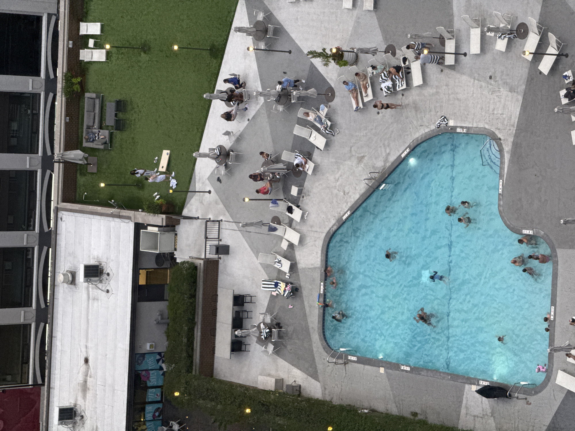 This aerial image captures a rooftop pool area and adjacent lounge space in Chicago, Illinois, USA. The scene is divided into distinct zones: a bright blue swimming pool in the foreground, and a green lawn area with seating and outdoor games in the background.
In the pool, several individuals are engaged in various water activities, including swimming and floating. The pool is surrounded by a concrete deck furnished with lounge chairs, some occupied by people relaxing.
Adjacent to the pool is a paved area with tables and chairs, and further back, a manicured lawn area. On the lawn, groups of people are seen socializing, some gathered around a game of cornhole, others lounging on sofas. Umbrellas are scattered throughout the outdoor spaces, providing shade. In the background, a building with dark-windowed facades looms over the scene. The overall impression is one of a modern urban leisure space.