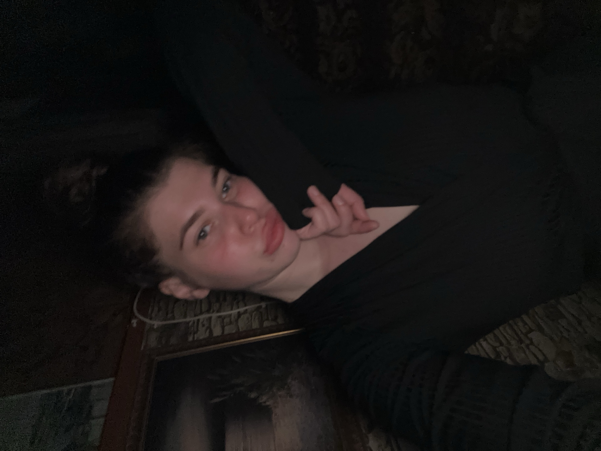 A young woman is lying down, wearing a black top and pants. She has her hair up in a bun. Her right hand is near her chin. The environment is dark, but what is visible looks to be an interior of a house. There appears to be a picture frame visible in the bottom left. No location in Мещёры, Russia can be confirmed.