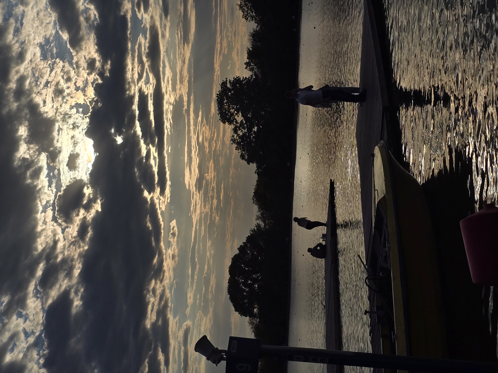 The image depicts a body of water, possibly a lake or river, under a sky with dramatic clouds and sunlight. In the distance, trees form a dark silhouette against the bright sky. A dock or pier extends into the water, with several people on it. One person stands near the end of the dock, while two other figures are smaller and appear to be further away. The water reflects the sunlight and clouds, creating a shimmering effect. In the foreground, part of a boat is visible, along with a post with signage. The image suggests a tranquil or recreational setting, with people enjoying the waterfront scenery. The location is Unknown, Unknown.