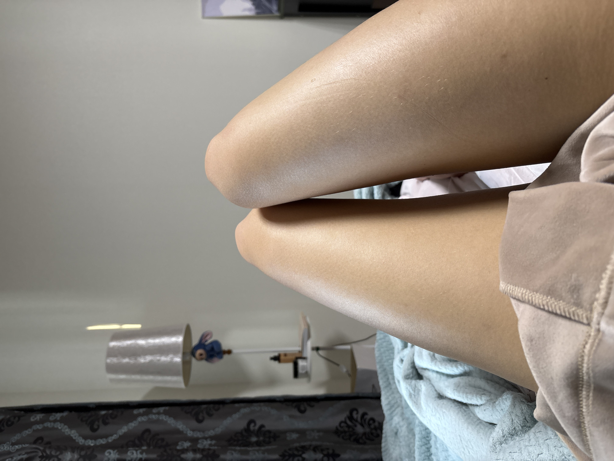 The image shows a close-up of a person's legs, specifically the knees, which are slightly bent and resting on each other. The skin tone is tan. Parts of the person's clothing are visible, including a tan-colored velvety garment and a blue towel underneath. In the background, there is a decorative object featuring a small blue bunny and a spool, as well as a bed with a patterned headboard. The image does not provide any information about the location or city of Ust-Ilimsk, Russia.