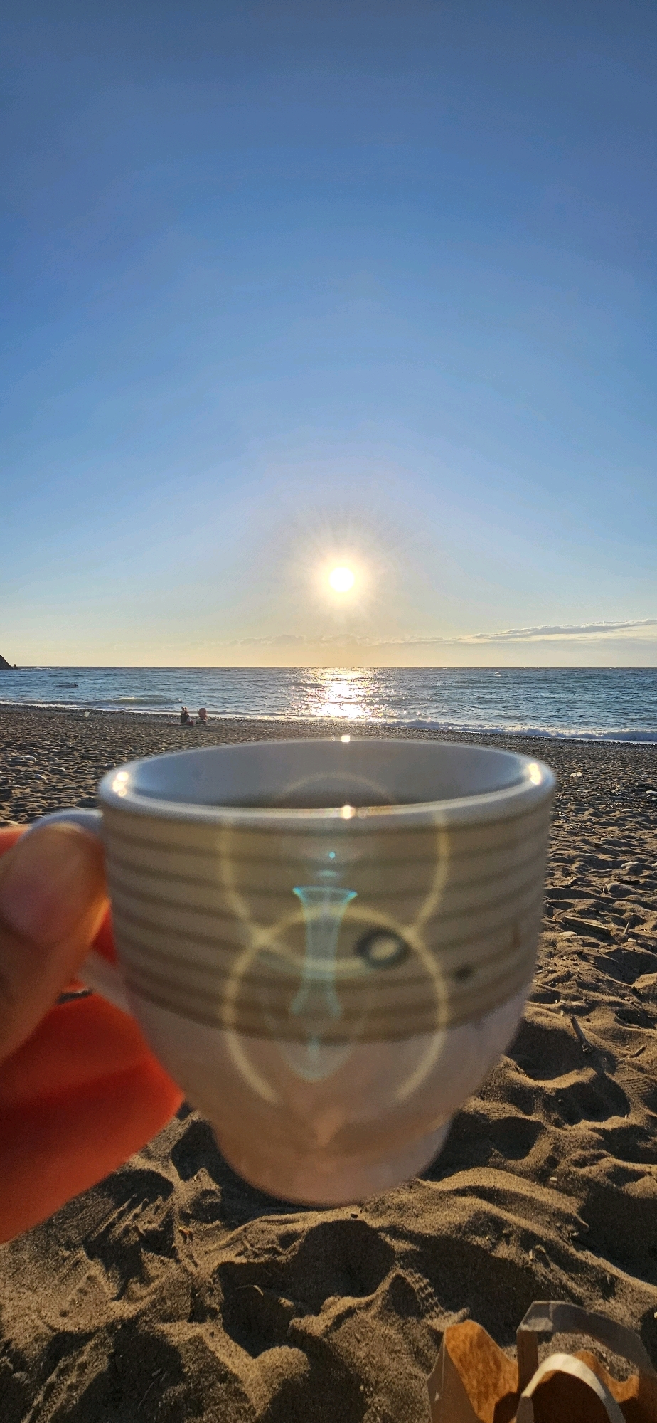 A close-up shot of a ceramic mug filled with a dark liquid, held by a person whose hand is visible in the lower left corner of the frame. The mug has a striped pattern. The background shows a beach scene with sand in the foreground and the sea and sky in the distance. The sun is setting or rising, casting a bright reflection on the water. Two silhouetted figures are visible sitting on the beach near the water's edge. The location is identified as the city of Kemer, Turkey. The overall impression is one of a serene morning or evening by the sea.