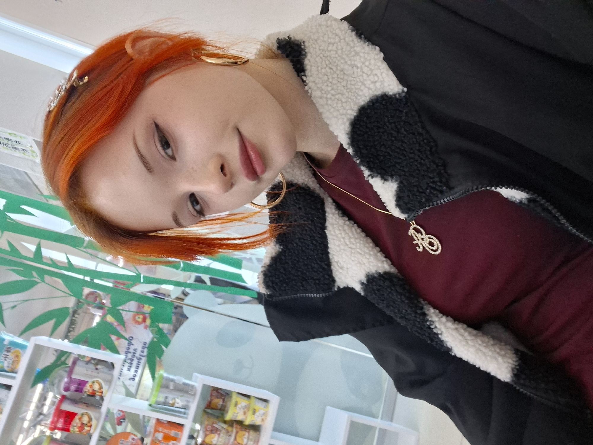A woman with orange hair stands indoors, wearing a burgundy top, a gold necklace with a pendant, a black jacket with a black and white faux fur collar, and gold hoop earrings. She has a decorative hair accessory and eyeliner. Shelves with products, decorations of bamboo, and a panda illustration are visible in the background, suggesting the location is a retail space in the city of Chaikovsky, Russia.