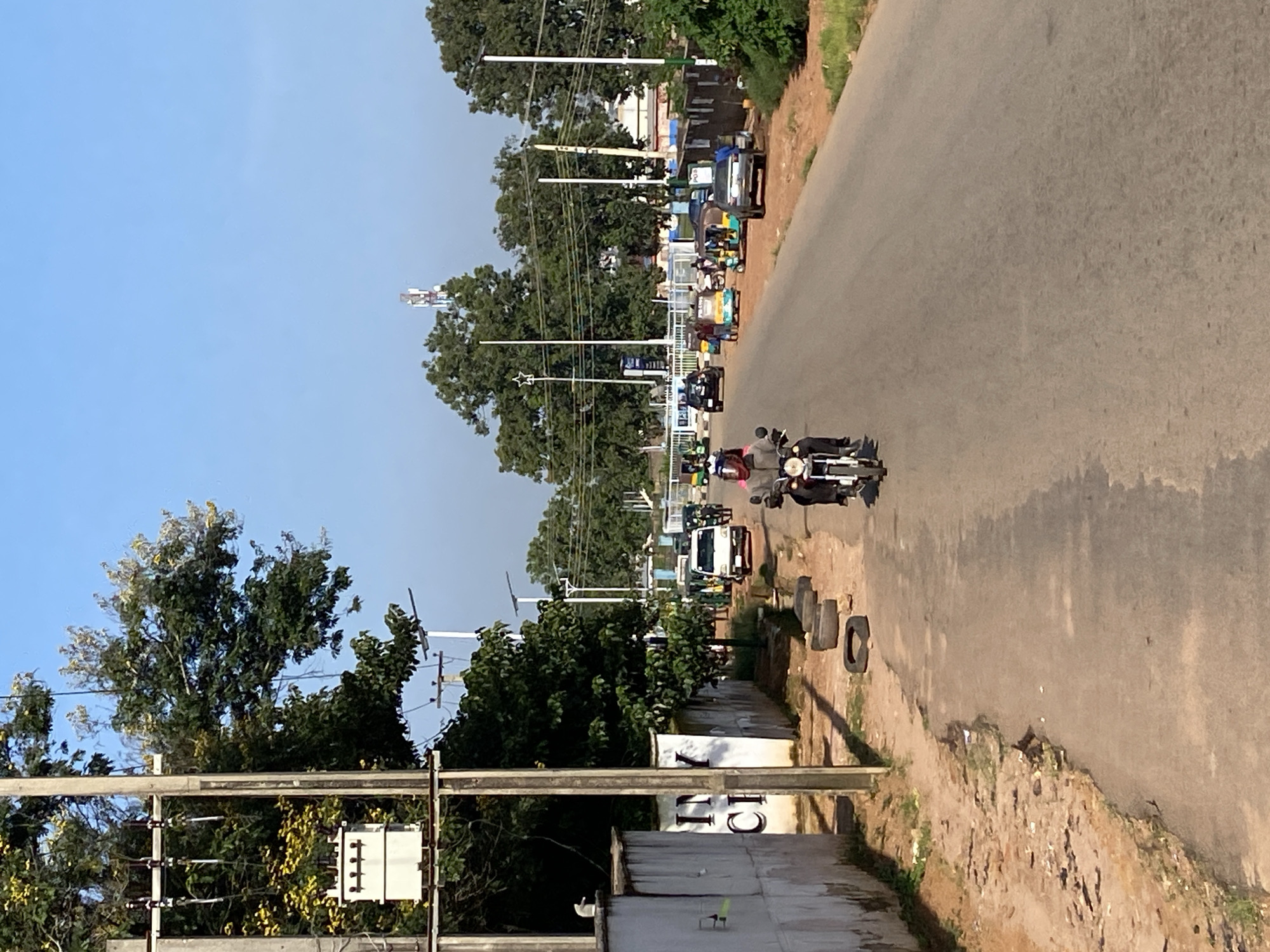 The image depicts a street scene in Gura Top, Nigeria. The road is paved and appears to be a main thoroughfare with multiple lanes. On the left side of the road, there is a white concrete wall with some faded text on it. Adjacent to the wall is a utility pole with electrical equipment, including a transformer, and trees with yellow flowers. Across the road, there are more trees, a communication tower in the distance, and several vehicles, including cars, minibuses, and auto-rickshaws, which are common in Nigeria.
In the foreground, a person is riding a motorcycle, wearing a helmet and a dark jacket. Several other vehicles and people are visible further down the road, indicating a moderate level of activity. The environment appears to be urban or semi-urban, with a clear blue sky above. The road is lined with some vegetation and there are discarded tires on the side of the road on the left. The overall impression is that of a typical day in a Nigerian city.