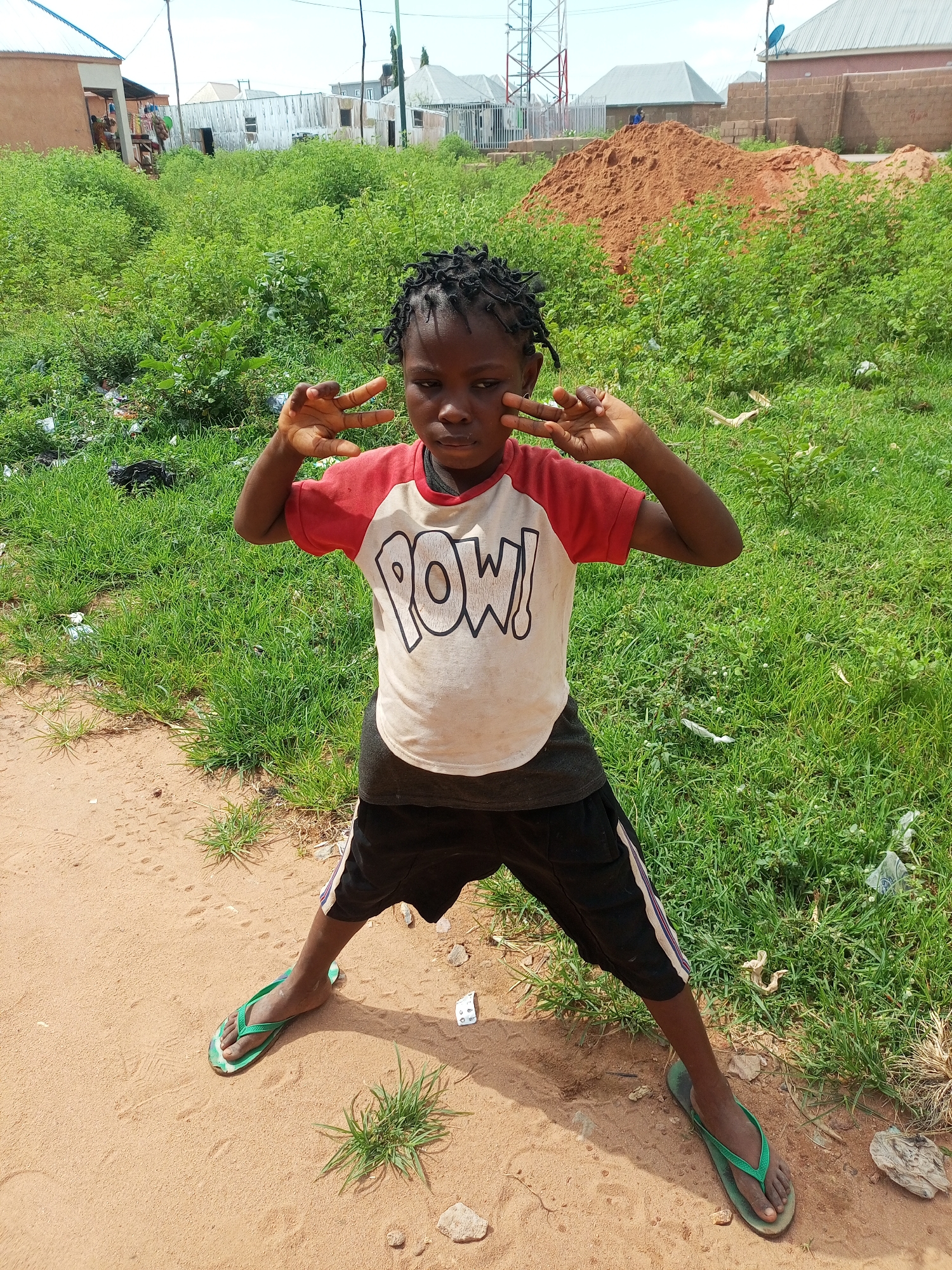 A young person stands in a dirt path in front of a grassy area, with buildings in the background. The individual, appearing to be a child, wears a red and white t-shirt with the word "POW!" printed on it, black shorts with white and purple stripes on the sides, and green flip-flops. They are holding both hands up, with index and middle fingers extended in a peace sign gesture, and looking towards the camera. The environment is dusty with sparse vegetation and several structures, including what appears to be a sand pile and a communication tower, suggesting a semi-urban or developing area in Tempure, Nigeria.