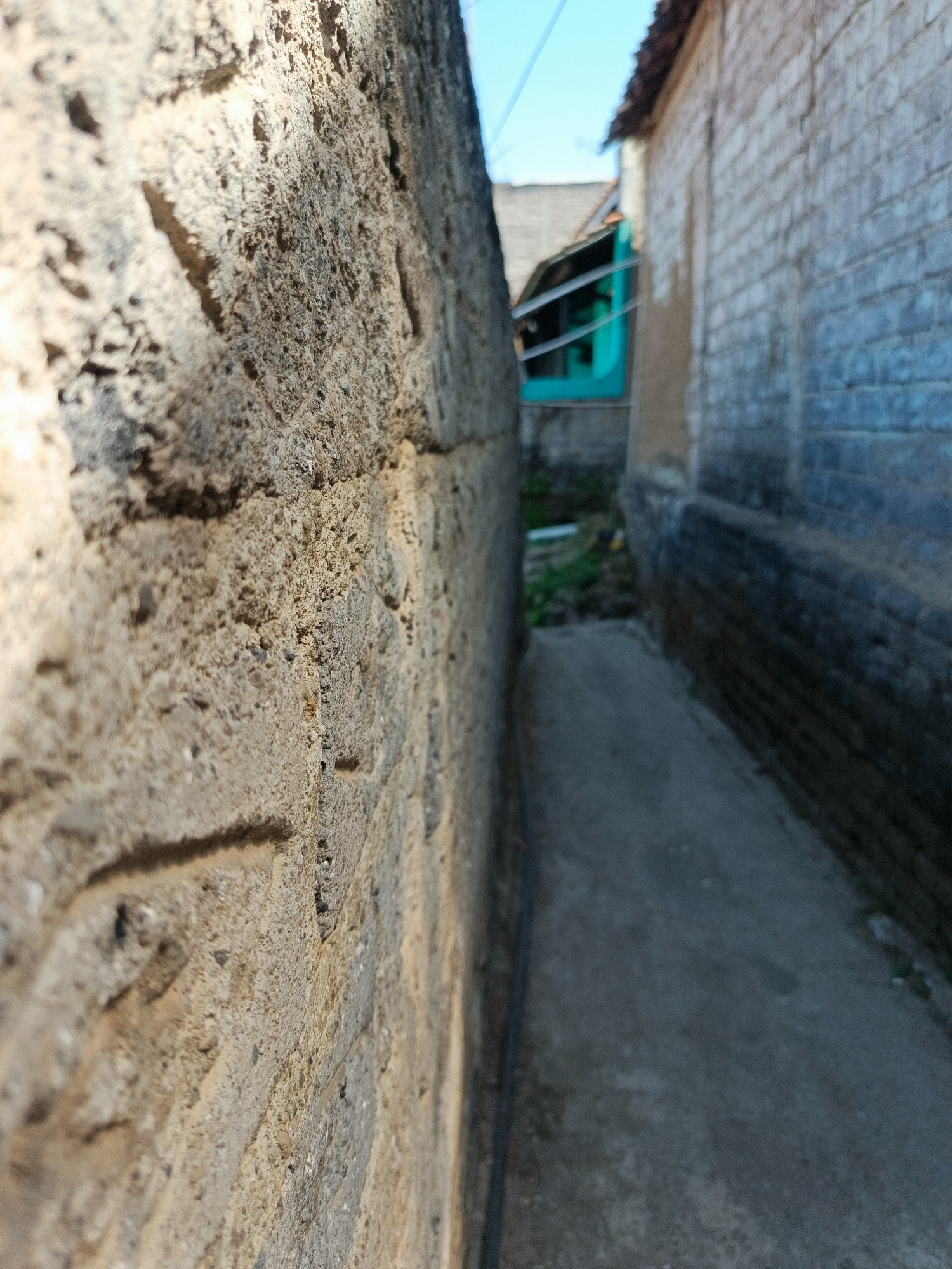 The image captures a narrow alleyway between two textured walls, likely located in Margajaya, Indonesia. The wall on the left is close to the camera and is made of rough, uneven stone or concrete, with visible shadows cast upon it by sunlight from the right. The wall on the right is constructed of light-colored bricks, with some darker areas suggesting wear or weathering. The alley itself is paved with a gray, slightly uneven surface, disappearing into the background. In the distance, a glimpse of buildings with tiled roofs and a bright teal-colored window or doorway can be seen. The sky above is a clear blue. No people or active events are depicted. The scene appears to be a quiet, everyday view of an urban environment.