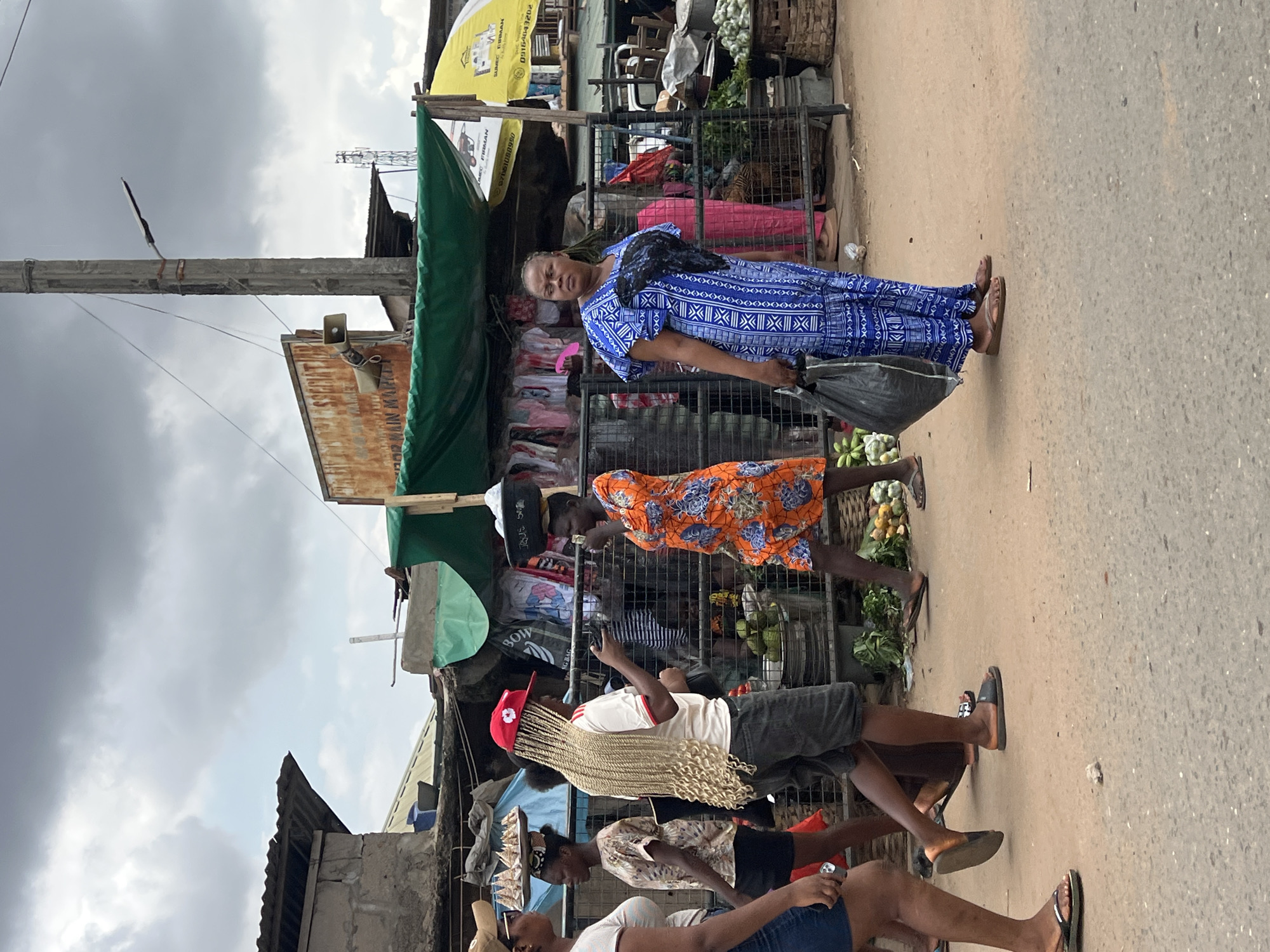 The image depicts a street scene in Nkpor, Nigeria, under a cloudy sky. In the foreground, a woman wearing a blue and white patterned dress and flip-flops walks with a black plastic bag. To her left, a person with long blonde braided hair, a red cap, and dark shorts is holding a phone. Behind them, a woman in an orange floral dress carries a basket on her head, and another woman in a patterned shirt and dark shorts walks to the left. The background features market stalls with green and yellow awnings, displaying clothing and produce. Various people are visible within the market area. The ground is a paved street.