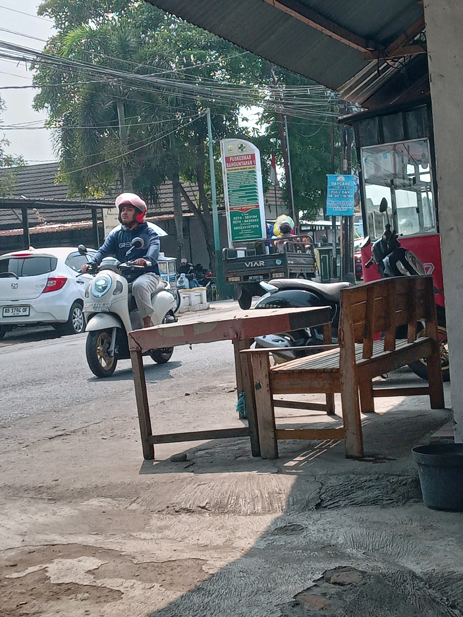 A person wearing a pink helmet and a blue jacket rides a white scooter down a paved road in Banguntapan, Indonesia. The scooter has a license plate that reads "AB 5966 UU". A white car with a license plate "AB 1794 DF" is parked on the side of the road to the left of the scooter. In the background, there are trees, a building with a corrugated metal roof, and signs. One sign reads "PUSKESMAS BANGUNTAPAN 1". To the right, there is a wooden bench and a dark plastic pot. The scene is outdoors, with sunlight casting shadows on the ground.
