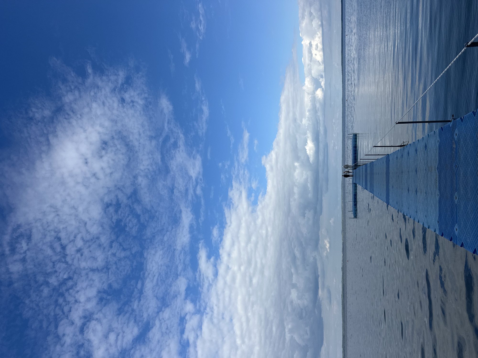 An elevated shot captures a blue plastic pontoon walkway extending into a calm, wide body of water under a vast sky filled with scattered white and grey clouds. The water surface exhibits ripples, reflecting the sky's blues and whites. Several figures, appearing as small silhouettes, are positioned on the walkway. In the distance, a faint treeline is visible across the water, suggesting land. The scene is tranquil and open, with the pontoon walkway leading the eye towards the horizon. The location is identified as Нарацкі сельскі Савет, Belarus.