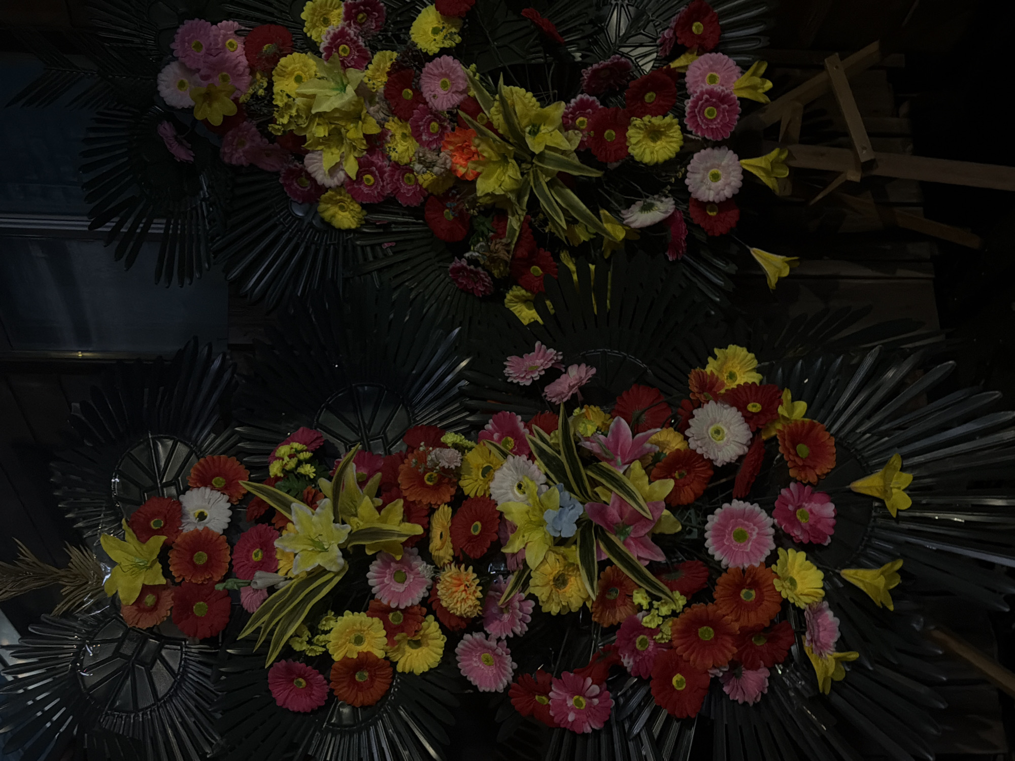 The image depicts two elaborate floral arrangements displayed on dark, fan-shaped backdrops. The flowers are a vibrant mix of colors, including red, pink, yellow, orange, and white, and appear to be artificial. The arrangement on the left is larger and more centrally positioned, while the one on the right is slightly smaller and to the right. Both arrangements feature a variety of flower types, with some appearing to be gerbera daisies and lilies. Some yellow, bell-shaped flowers hang down from the bottom of the arrangements. The backdrop of the flowers on the left is circular with radiating lines, while the backdrop on the right has a similar radiating design but is less distinctly circular. The arrangements are set against a dark background, possibly an indoor space with low lighting, and a portion of a wooden easel is visible beneath the right-hand arrangement. The scene is consistent with a decorative display, possibly for an event or establishment in Dongducheon-si, South Korea.