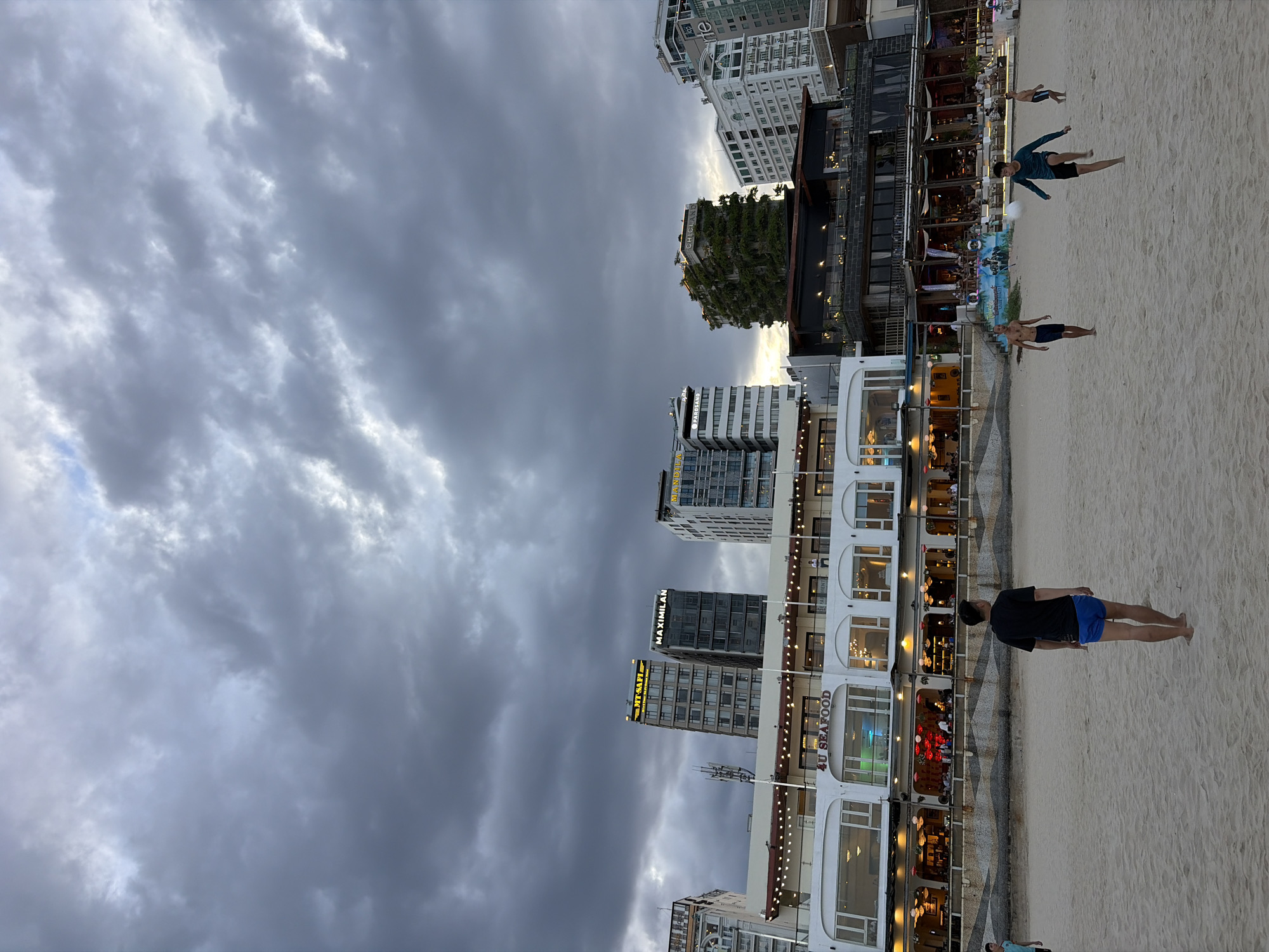 This image depicts a cloudy day on a sandy beach in Đà Nẵng, Vietnam. In the foreground, several individuals are visible on the beach. A man in a black t-shirt and blue shorts is walking away from the camera. Further down the beach, three young men are playing. One is kicking a ball, another is running, and a third is standing.
In the background, a row of buildings lines the beachfront. The closest building appears to be a multi-story restaurant or establishment with large windows and outdoor seating. Signage on this building reads "4U SEAFOOD." Behind this, a collection of taller buildings, including skyscrapers with distinctive architecture, dominates the skyline. One building is notably covered in dense greenery. The sky is filled with heavy, dark gray clouds, suggesting overcast weather or an approaching storm. The overall scene captures a moment of leisure on a coastal urban landscape.