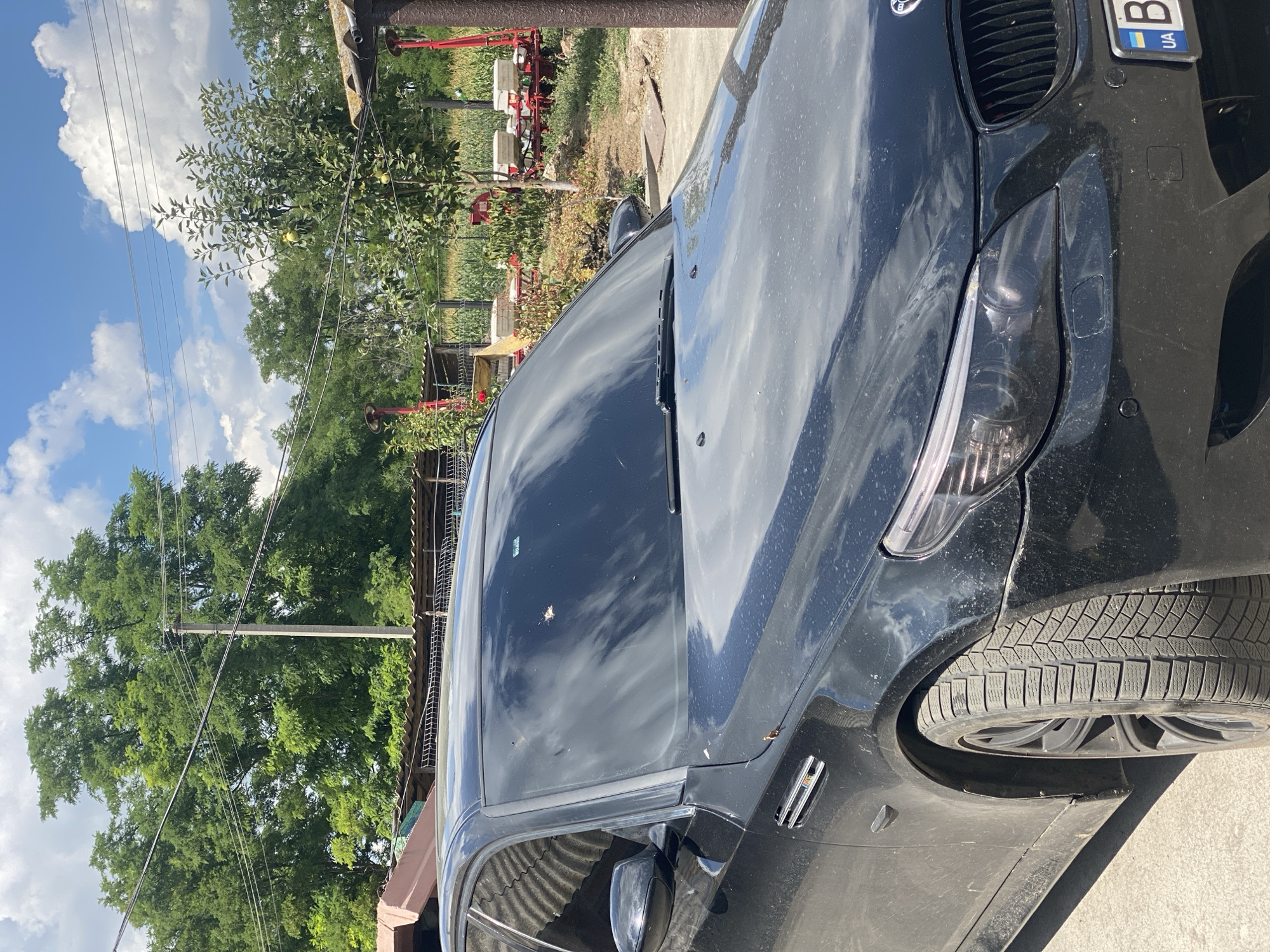 The image shows a black BMW sedan parked outdoors, likely in a rural or semi-rural setting given the background of trees, a field, and what appears to be agricultural equipment. The car's windshield is visible, reflecting the cloudy sky. In the background, there are power lines and utility poles, suggesting the presence of infrastructure. Further back, there's a cornfield, some bushes, and what looks like a simple structure or shed. The location is identified as Pletenyi Tashlyk, Ukraine. There are no people visible in the image. The car is parked on a paved or dirt surface.