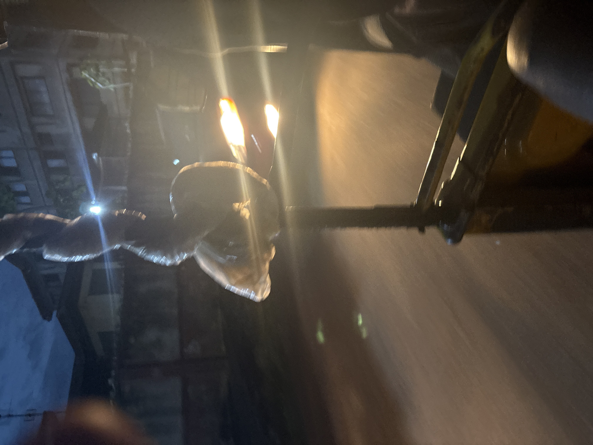 This is a nighttime, blurry photograph taken from inside a moving vehicle, likely a rickshaw or a similar open-air transport, in Onitsha, Nigeria. The foreground shows parts of the vehicle's interior, including yellow and dark-colored seats and metal bars. In the mid-ground, a person is holding a clear plastic bag containing an object, possibly food, illuminated by a bright light source. There are also bright, blurred lights in the background, indicative of streetlights or vehicle headlights. The environment appears to be an urban street with buildings and a fence visible. The overall impression is a candid shot capturing a moment of transit at night.