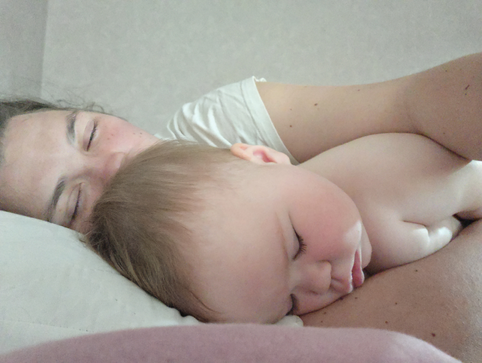 The image depicts an adult and a baby sleeping closely together on a bed. The adult is lying on their back with their eyes closed, and is wearing a white shirt. The baby is lying on their side, nestled against the adult, also with eyes closed and tongue slightly visible. They are not wearing any clothes. The background consists of a white pillow and a light gray wall.
The location in the city of Chaikovsky, Russia, is not determinable from the image.